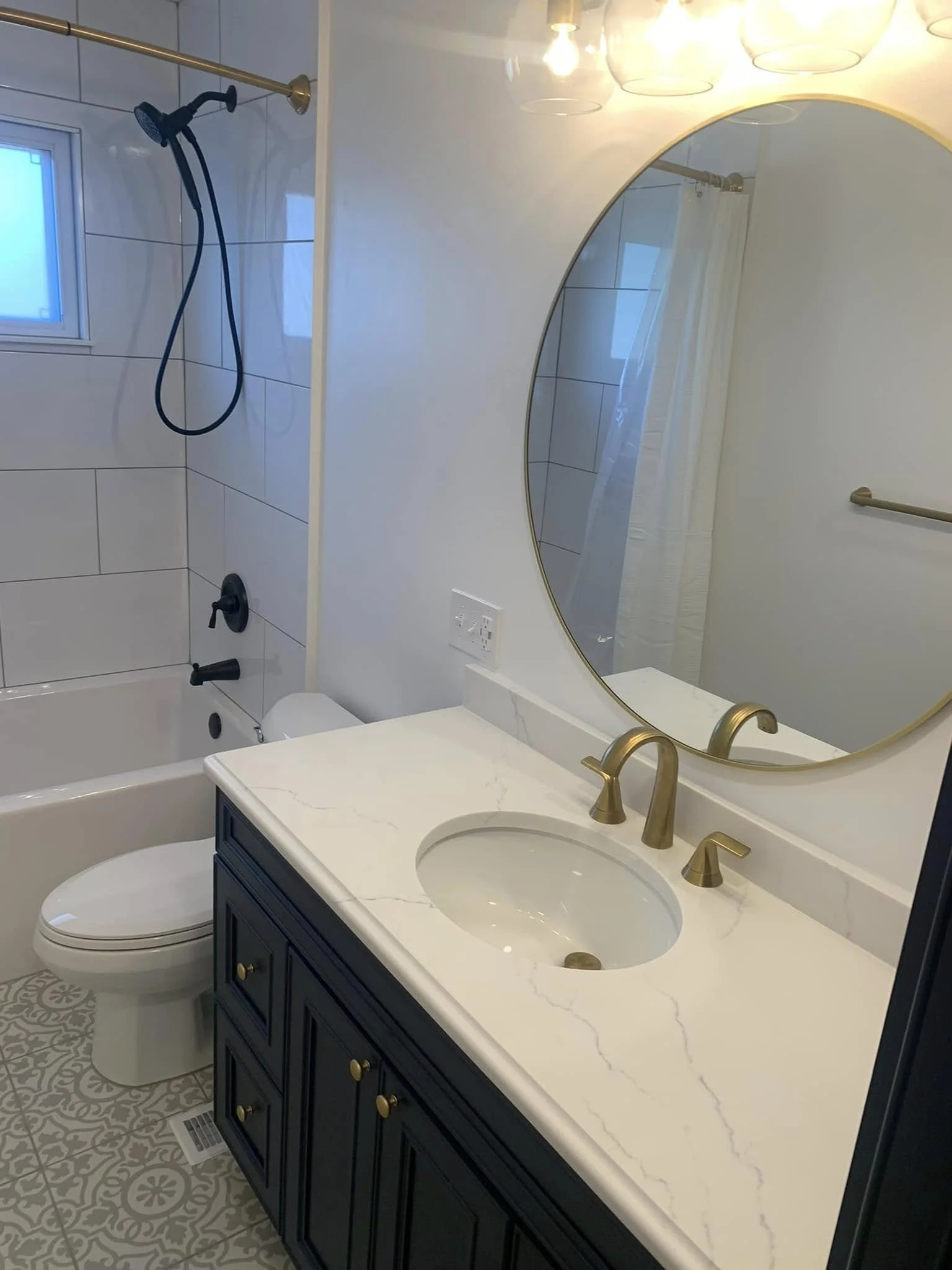 Bathroom with a dark blue vanity, white marble countertop with gold fixtures, a round mirror, a bathtub with a black showerhead, a window, and tiled walls.