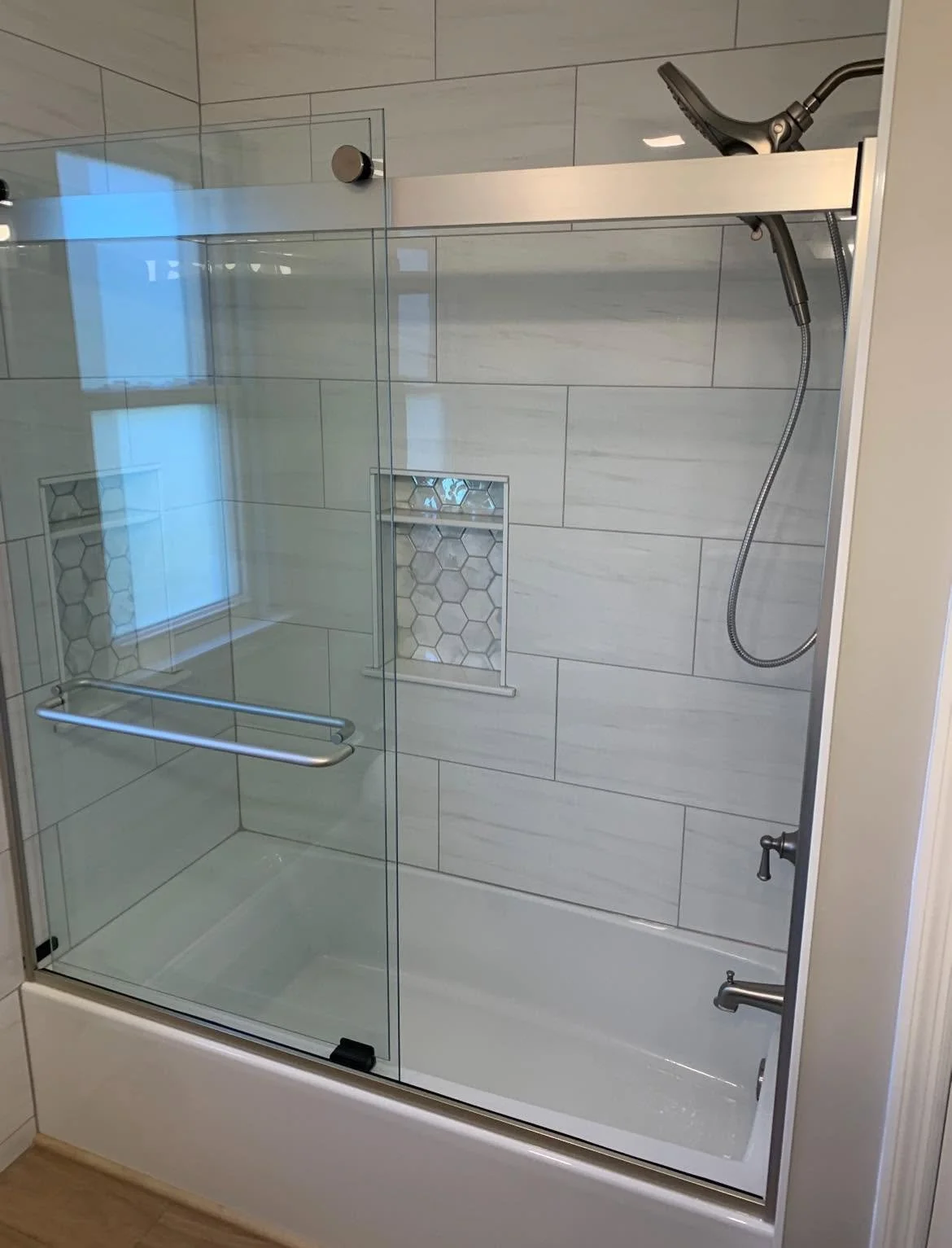 A modern walk-in shower with glass doors, a handheld showerhead, a built-in niche with hexagonal tiles, and a grab bar, surrounded by large white tiles.