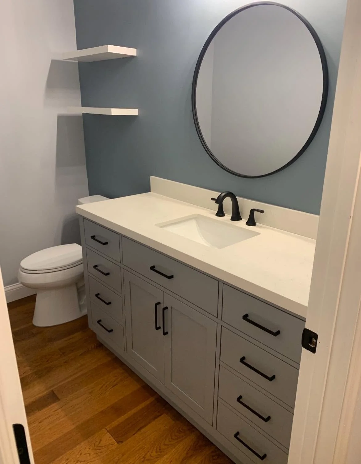Modern bathroom with a gray vanity, black hardware, a white countertop, an oval mirror, a black faucet, and a toilet, with light blue walls and wooden flooring.