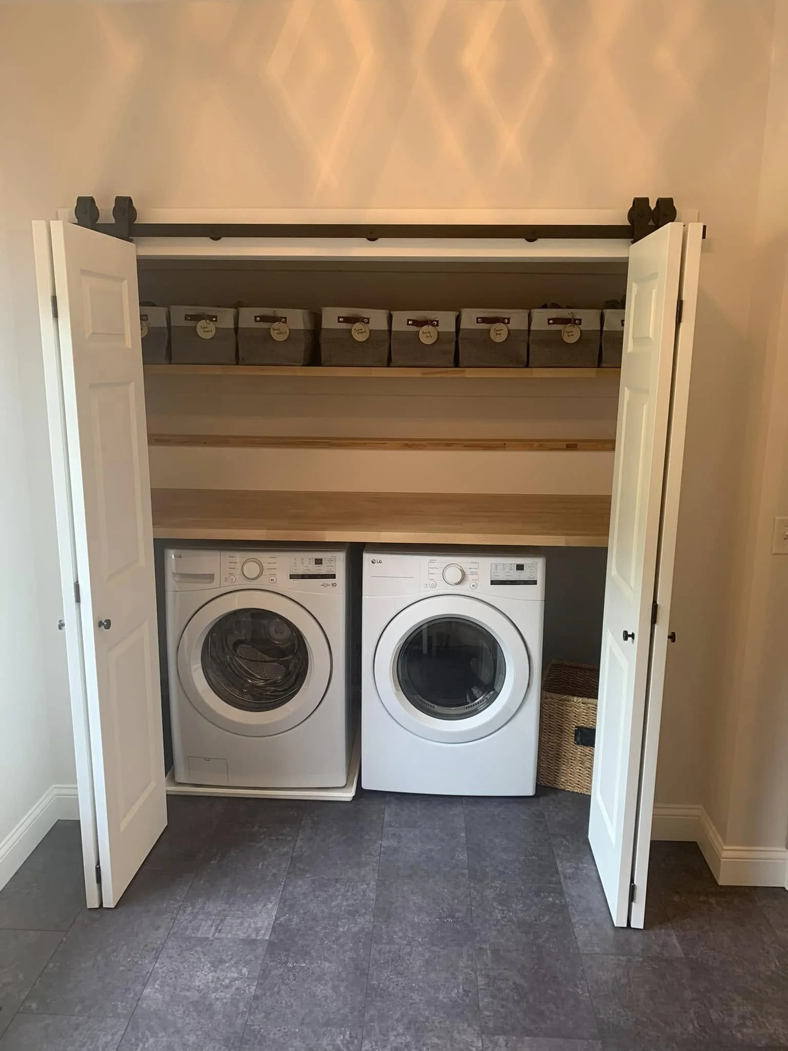 A laundry closet with two white front-loading washing machines side by side. Above the machines is a wooden shelf with six fabric bins labeled with tags. The closet has open double doors, beige walls, and gray floor tiles.