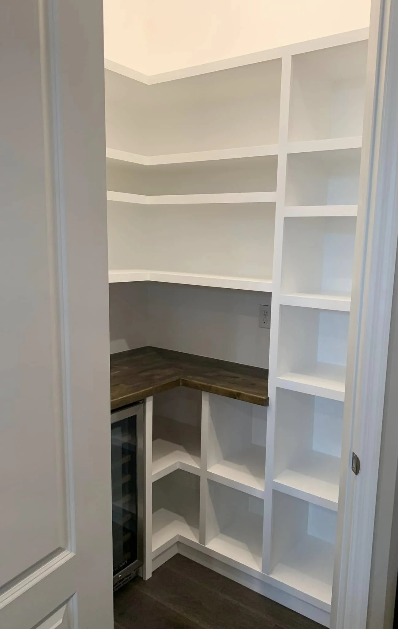 Empty white built-in shelving unit in a corner of a room with a dark wooden countertop and a small wine fridge below.
