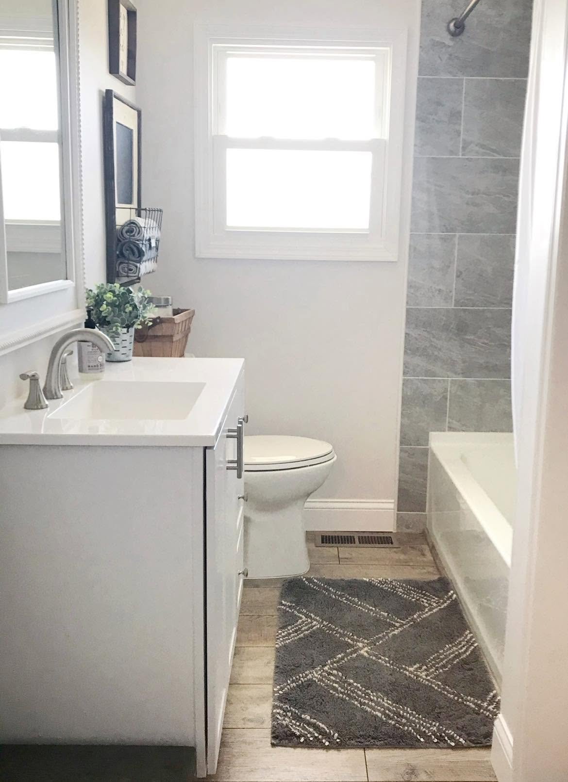 A modern small bathroom with a white vanity, a toilet, a gray tiled shower, a window, and a decorative gray rug on light wood flooring.