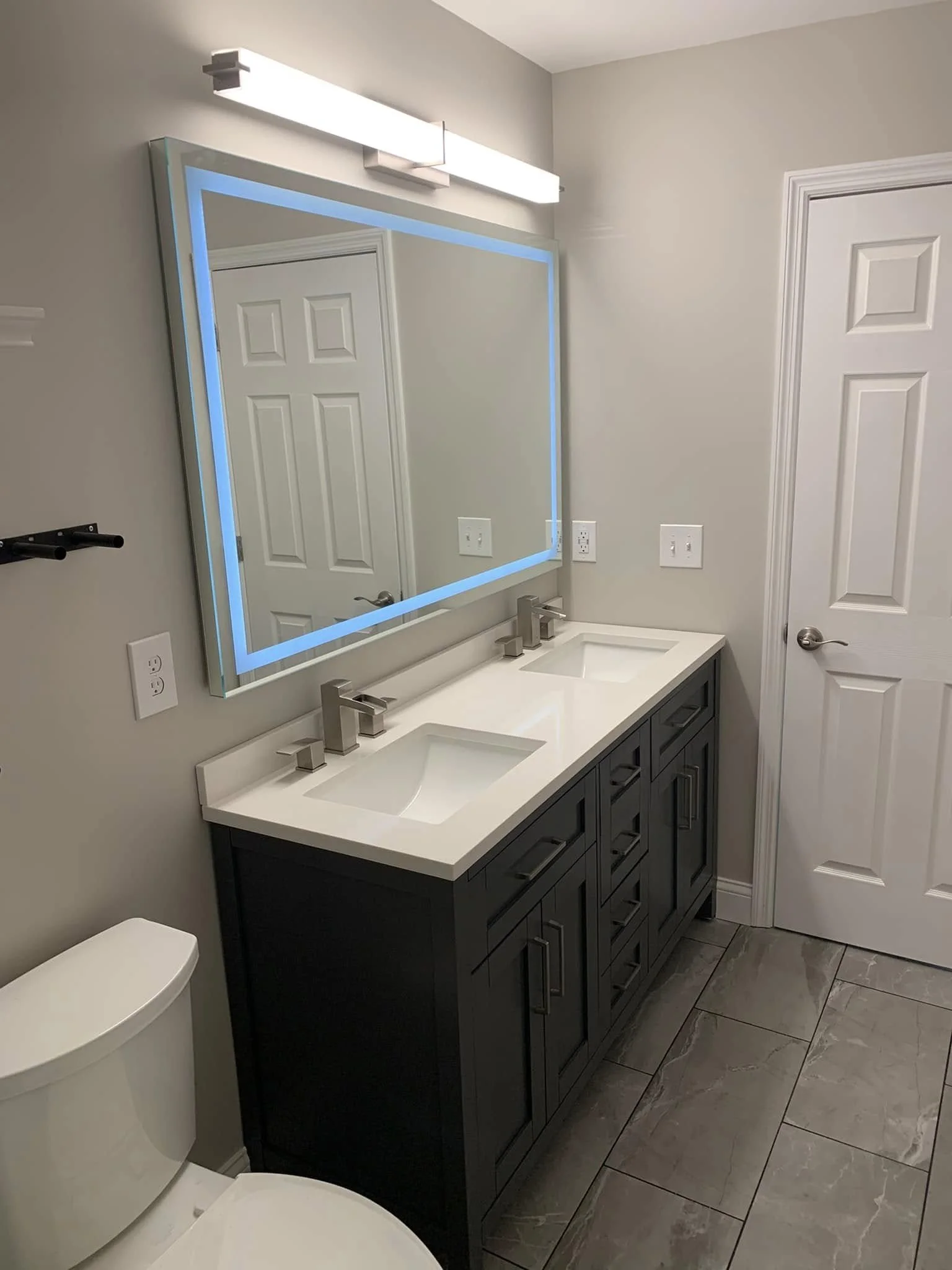 Bathroom with a double vanity, illuminated mirror with LED lighting, white countertop, and gray cabinetry, next to a toilet and a closed door.