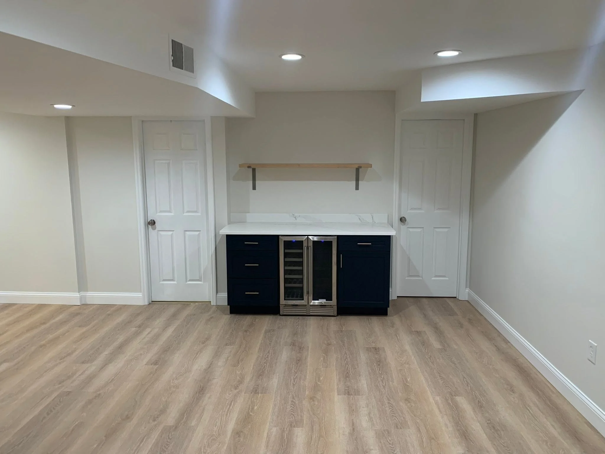 Empty basement or entertainment room with white walls, hardwood floors, ceiling lights, two white doors, a black cabinet with a built-in wine cooler, and a wooden shelf on the wall.