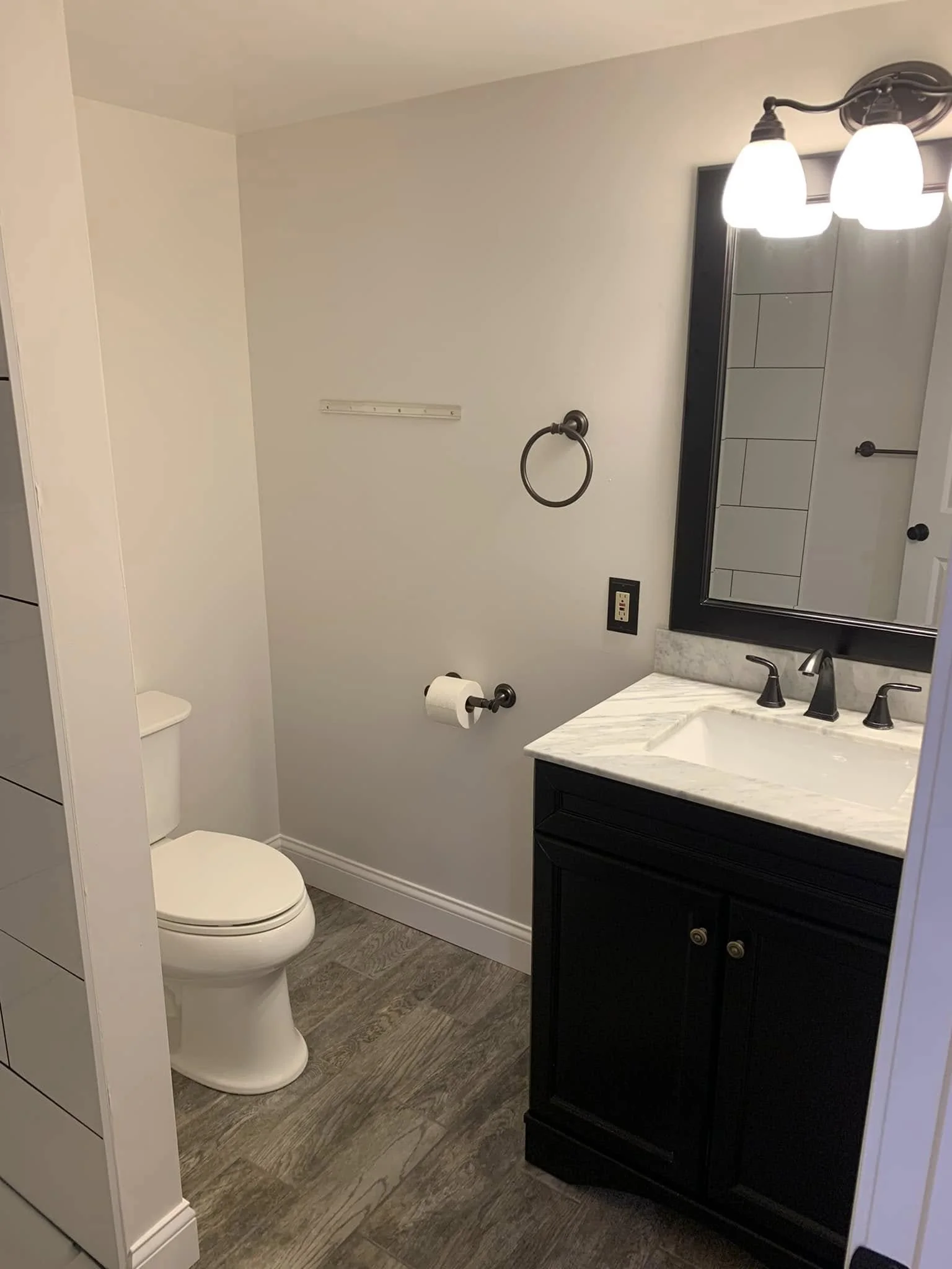 A small bathroom with a toilet, a black vanity with a marble countertop, a mirror, and a light fixture with two bulbs.
