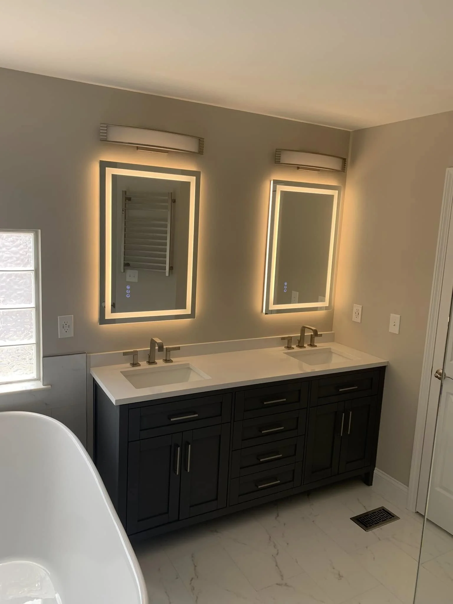 Modern bathroom with a white dual-sink vanity, black cabinetry, illuminated rectangular mirrors, and beige walls. A window with textured glass and a floor vent are visible.