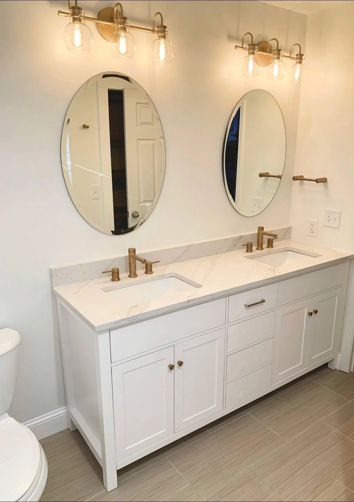 Double bathroom vanity with white marble countertop, two oval mirrors, and brass fixtures, illuminated by two brass light fixtures with three bulbs each.