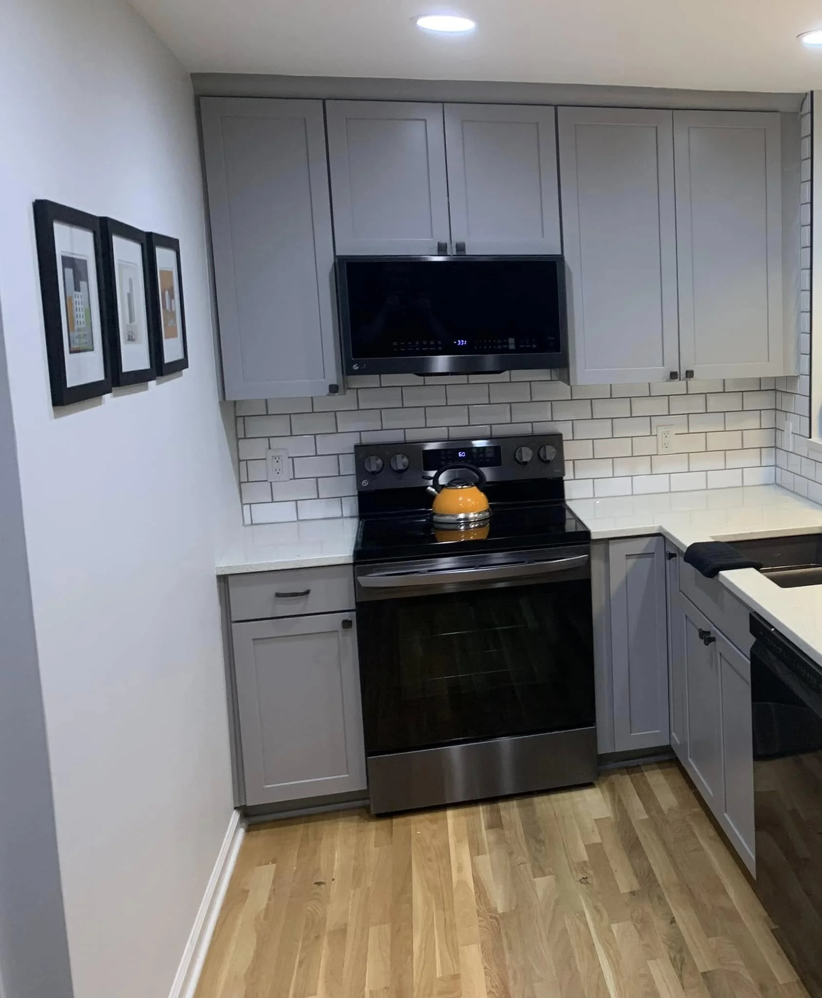 Kitchen with light gray cabinets, a black microwave, and a stainless steel stove with a yellow teapot on top. The counter is white, and the backsplash is made of white subway tiles. The floor has light wood planks, and three framed pictures hang on t