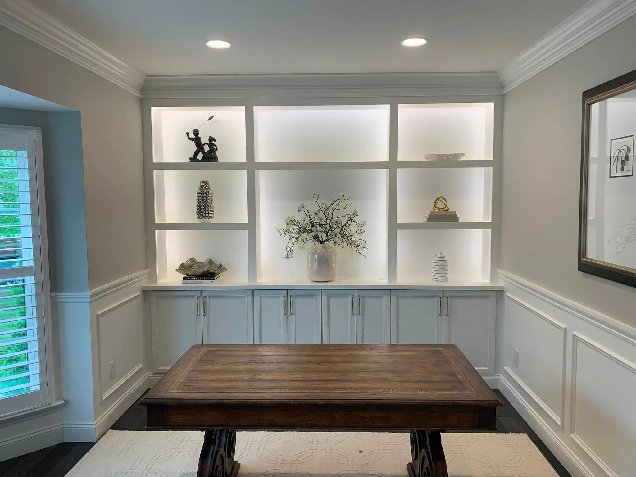 Built-in white shelving unit with decorative items including a sculpture, vase, and flowers, in a room with crown molding and hardwood flooring.