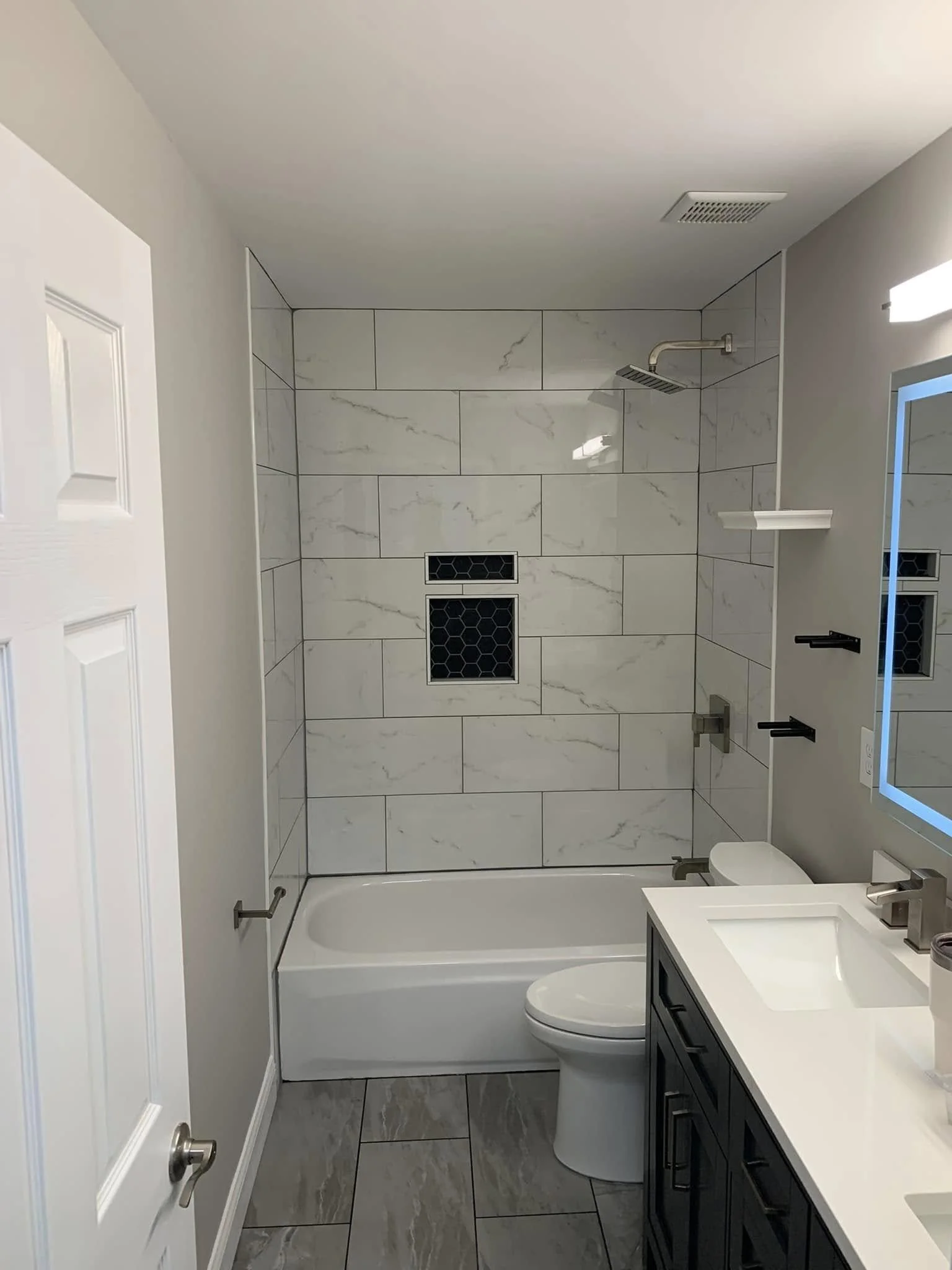 Modern bathroom with marble tiles on the wall and floor. Includes a white bathtub, a toilet, a dark vanity with a white countertop, and a mirror with a blue LED frame. Ceiling vent and showerhead visible.