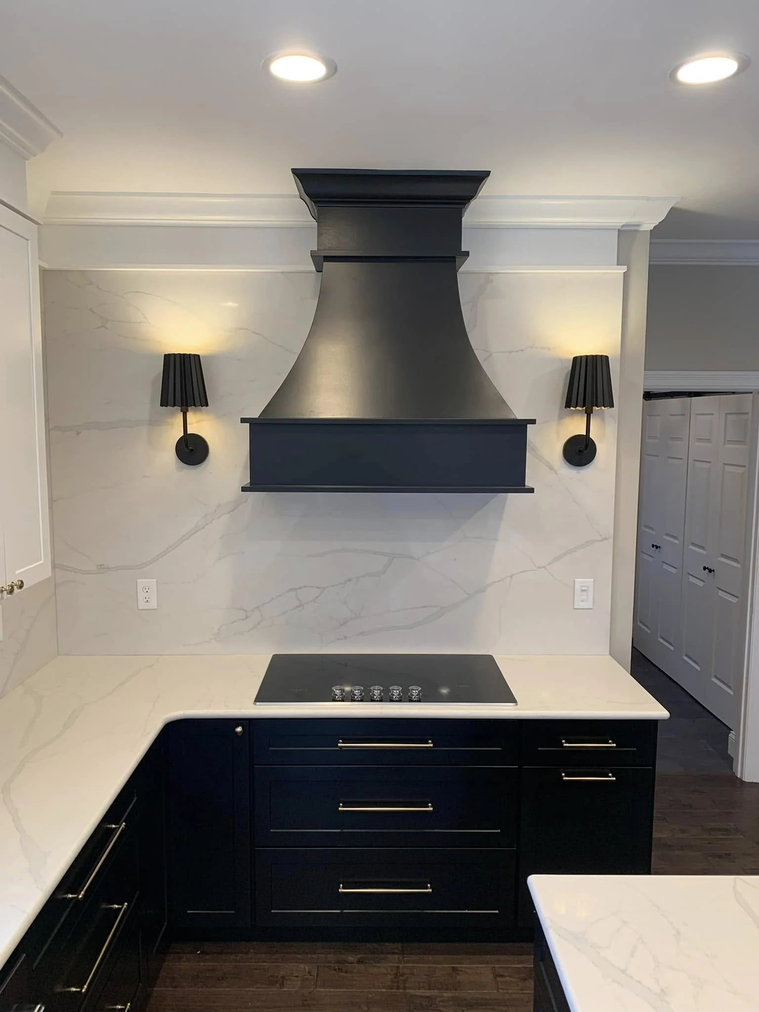 Modern kitchen with dark blue cabinets, a white marble countertop, a black range hood, wall-mounted black lamps, and a dark wood floor.