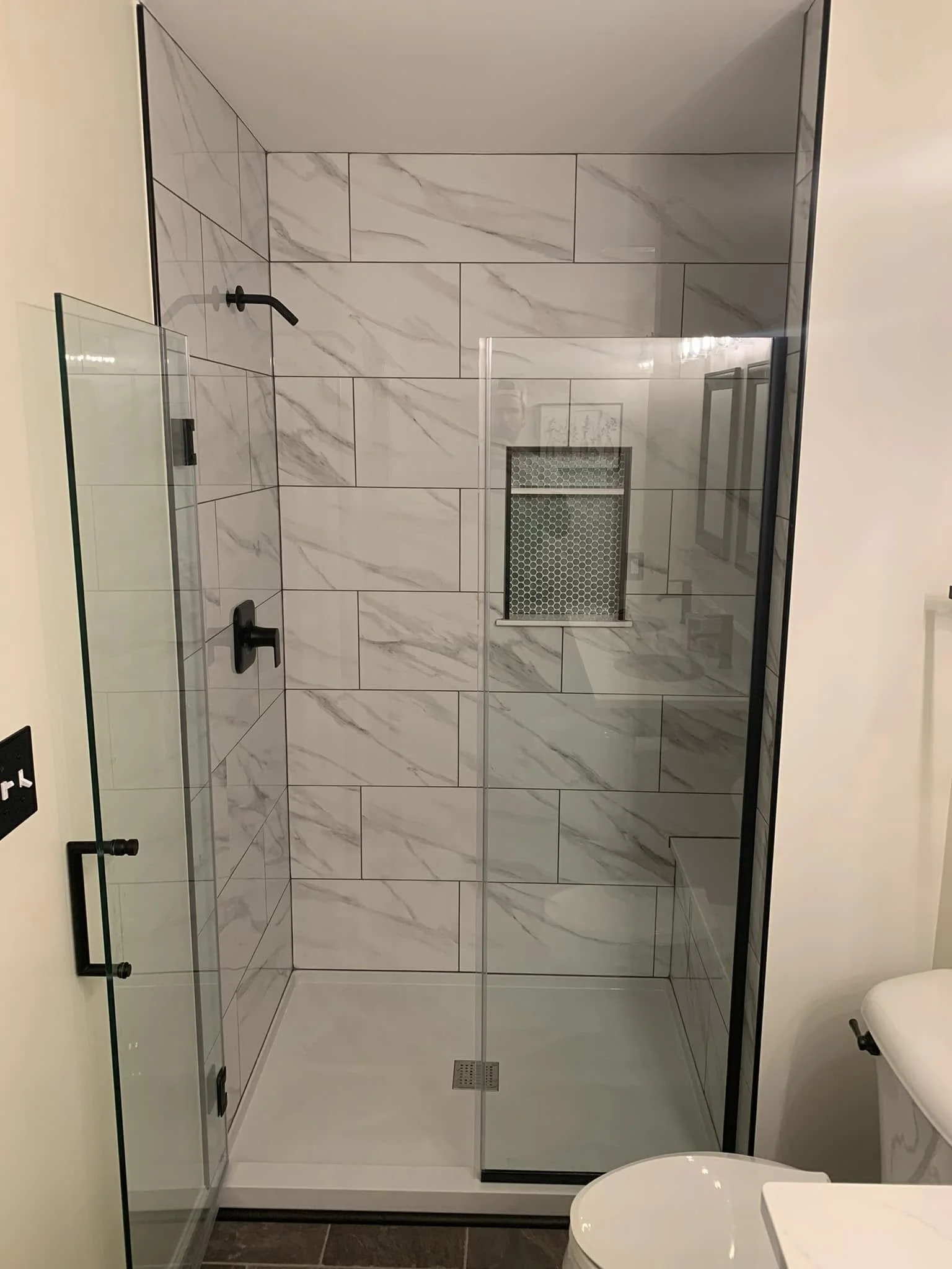 A modern walk-in shower with large white marble tiles, a black showerhead, and a small window with a hexagonal tile pattern, enclosed with a black-framed glass door.