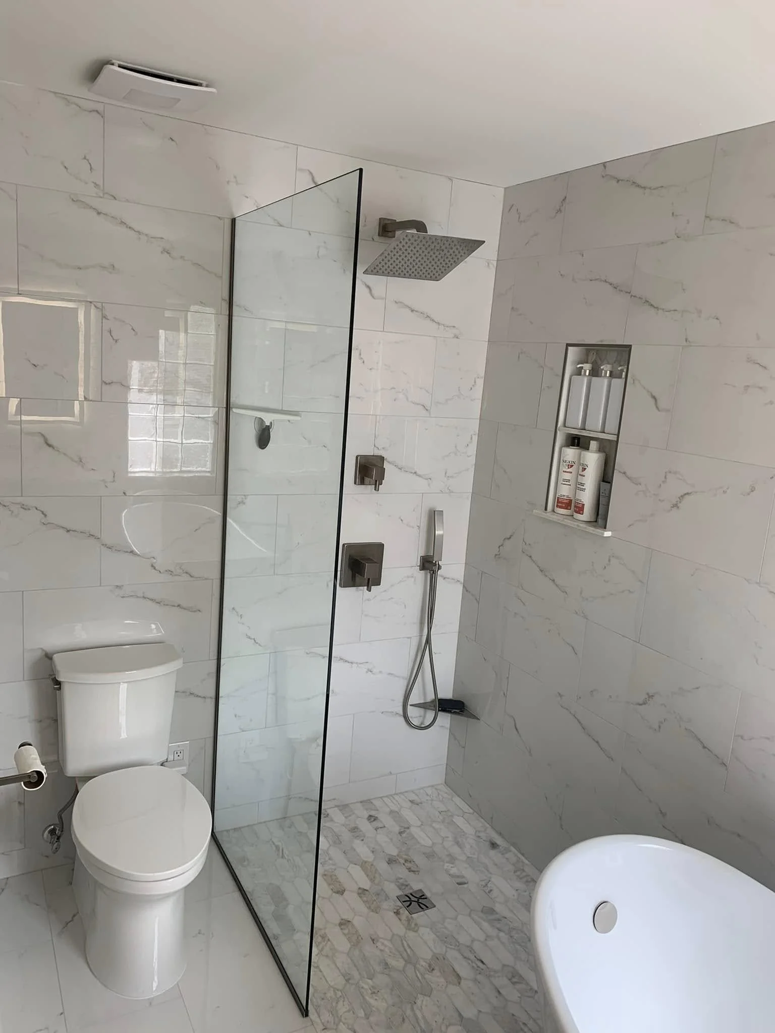 A modern bathroom with white marble tiles on the walls and floor. It features a toilet, a bathtub, a large shower with a glass partition, and a built-in wall shelf holding toiletries. The ceiling has a vent, and there is a window reflection visible o