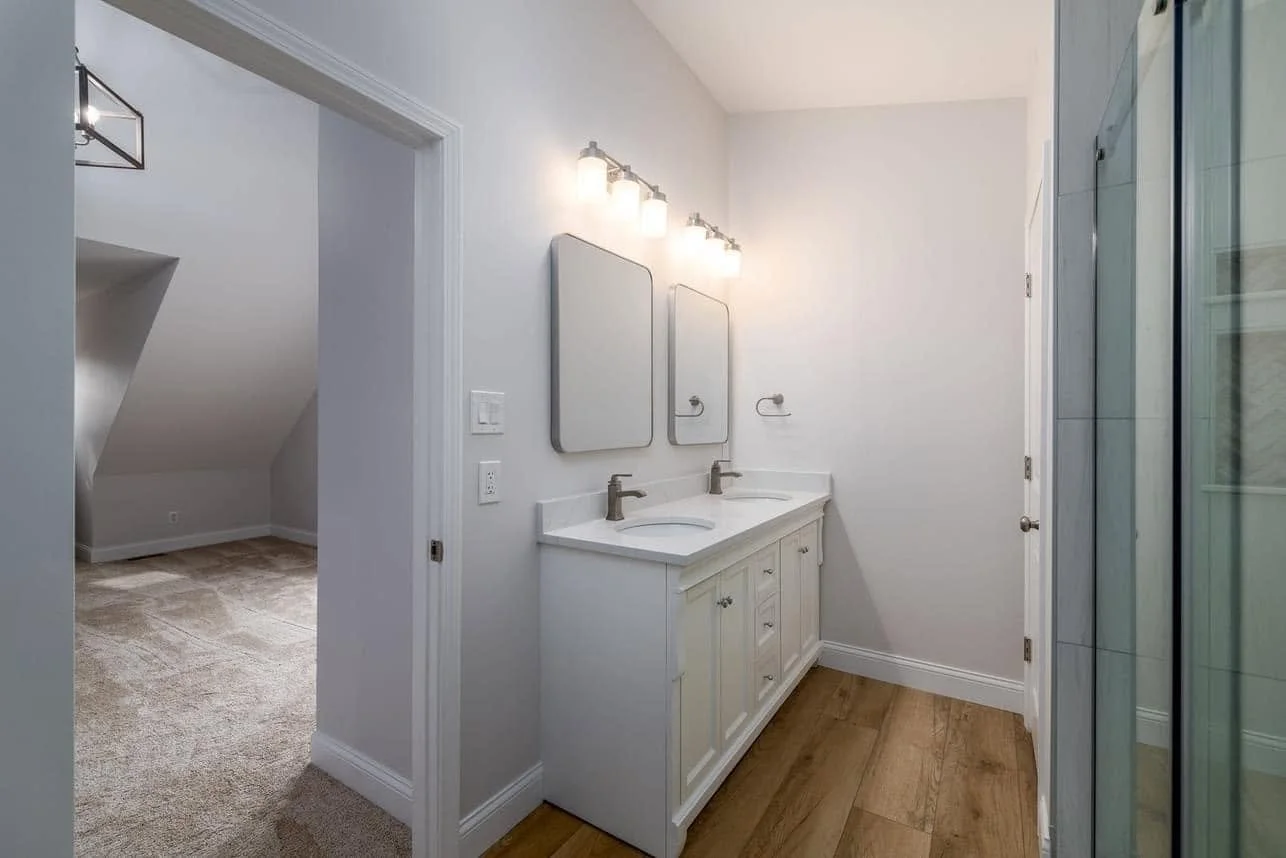 Bathroom with white vanity, twin sinks, and mirrors, overhead light fixtures, wooden floor, and glass shower enclosure on the right.