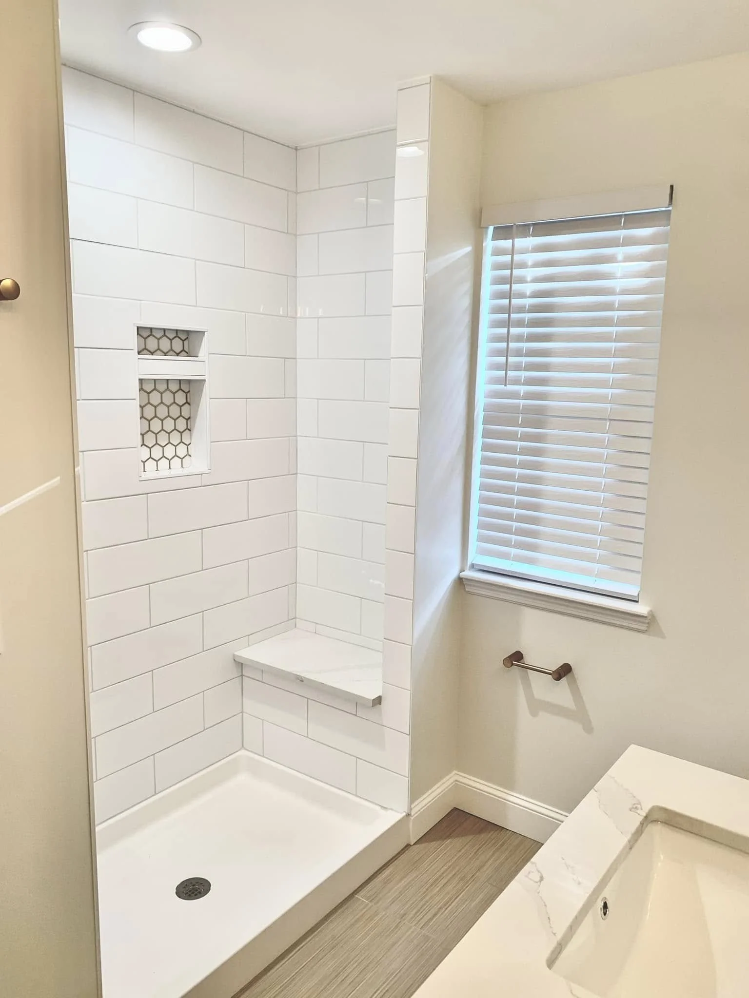 Bathroom with walk-in shower, white subway tiles, niche with hexagonal tiles, window with blinds, white marble countertop with sink.