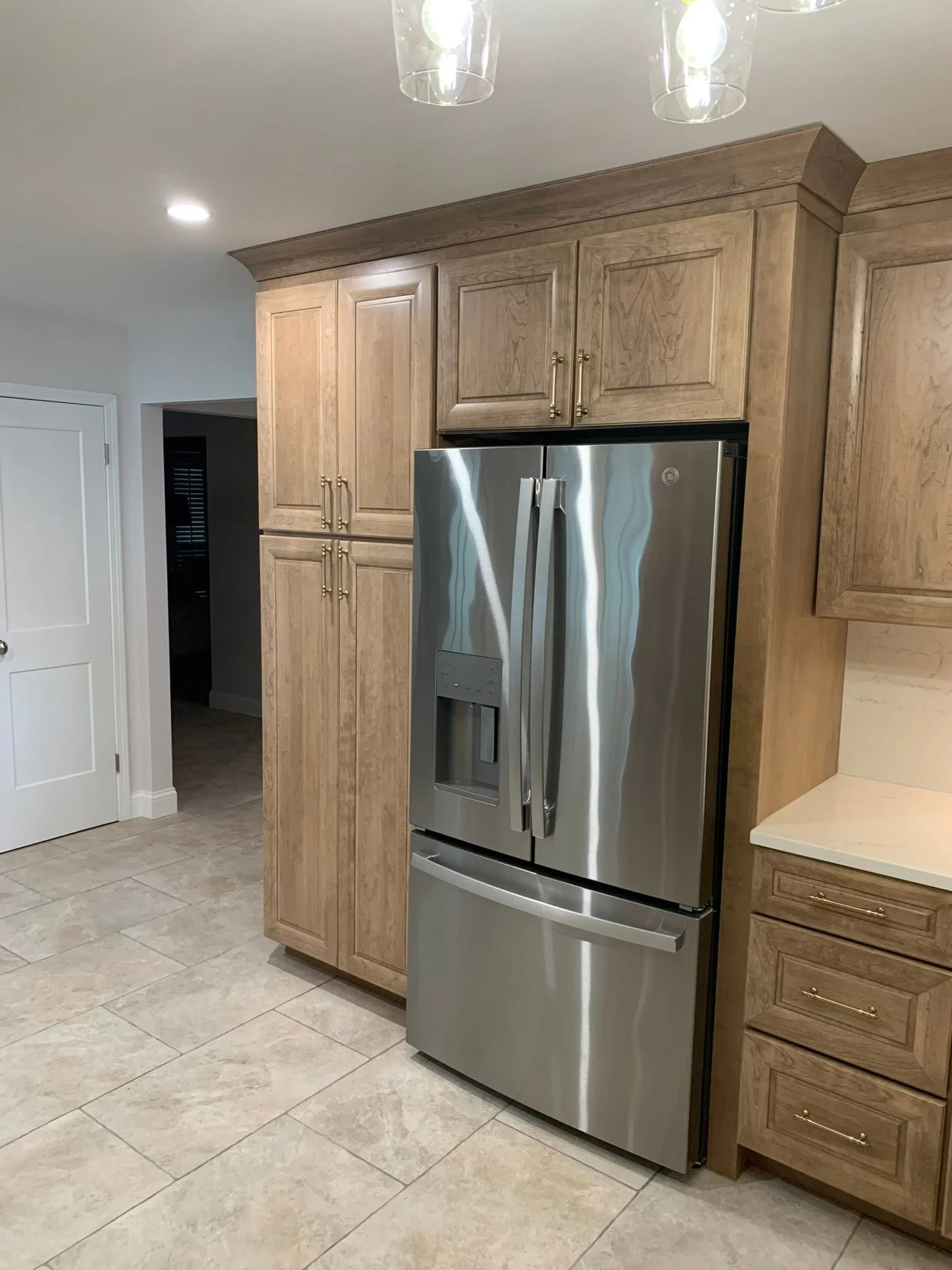A kitchen with wooden cabinets, a stainless steel refrigerator, beige tiled floor, and a white ceiling with recessed lighting and hanging glass pendant lights.