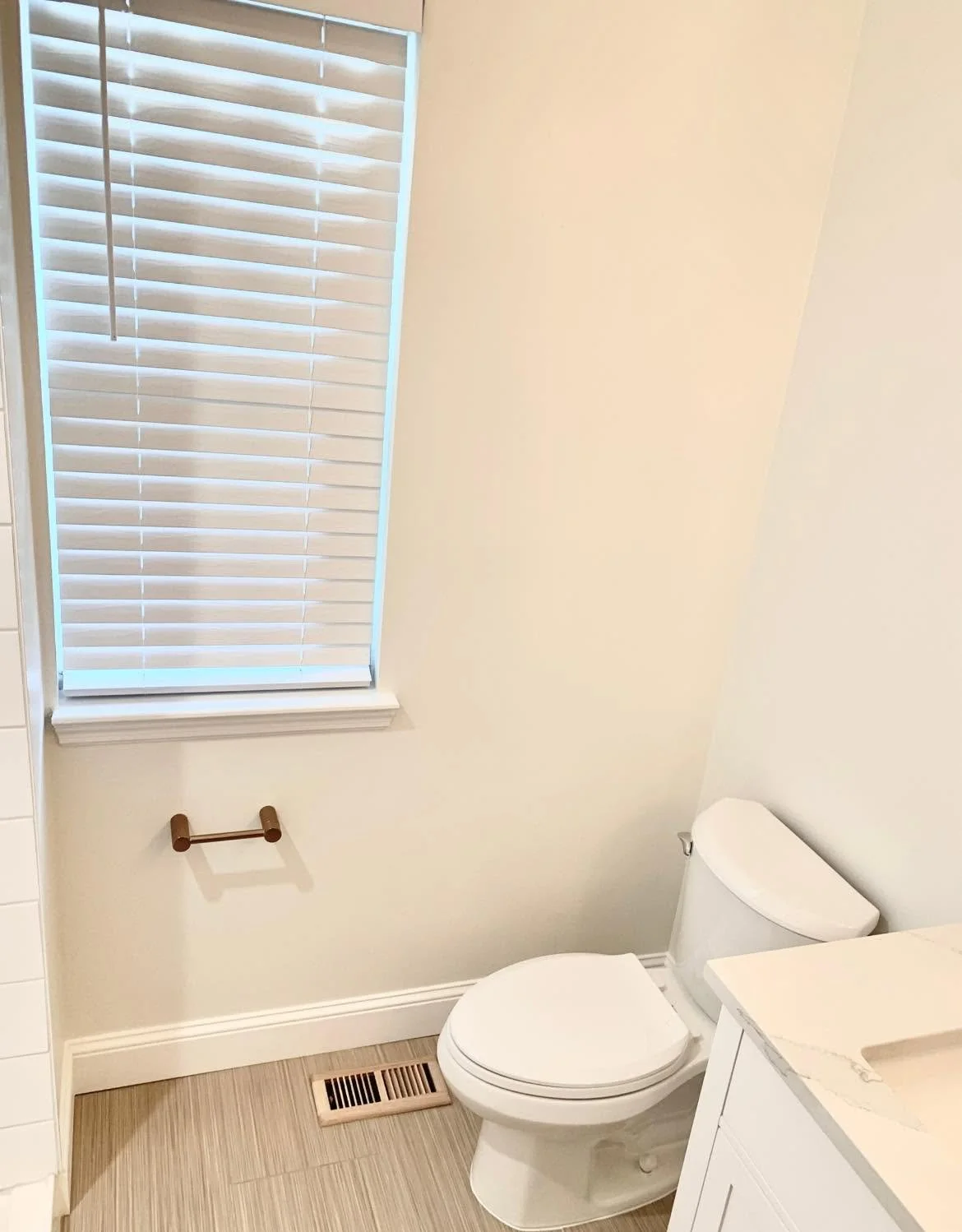 A small bathroom with a window with white blinds, a toilet, a white cabinet with a marble countertop, and a wood-colored toilet paper holder on the beige wall.