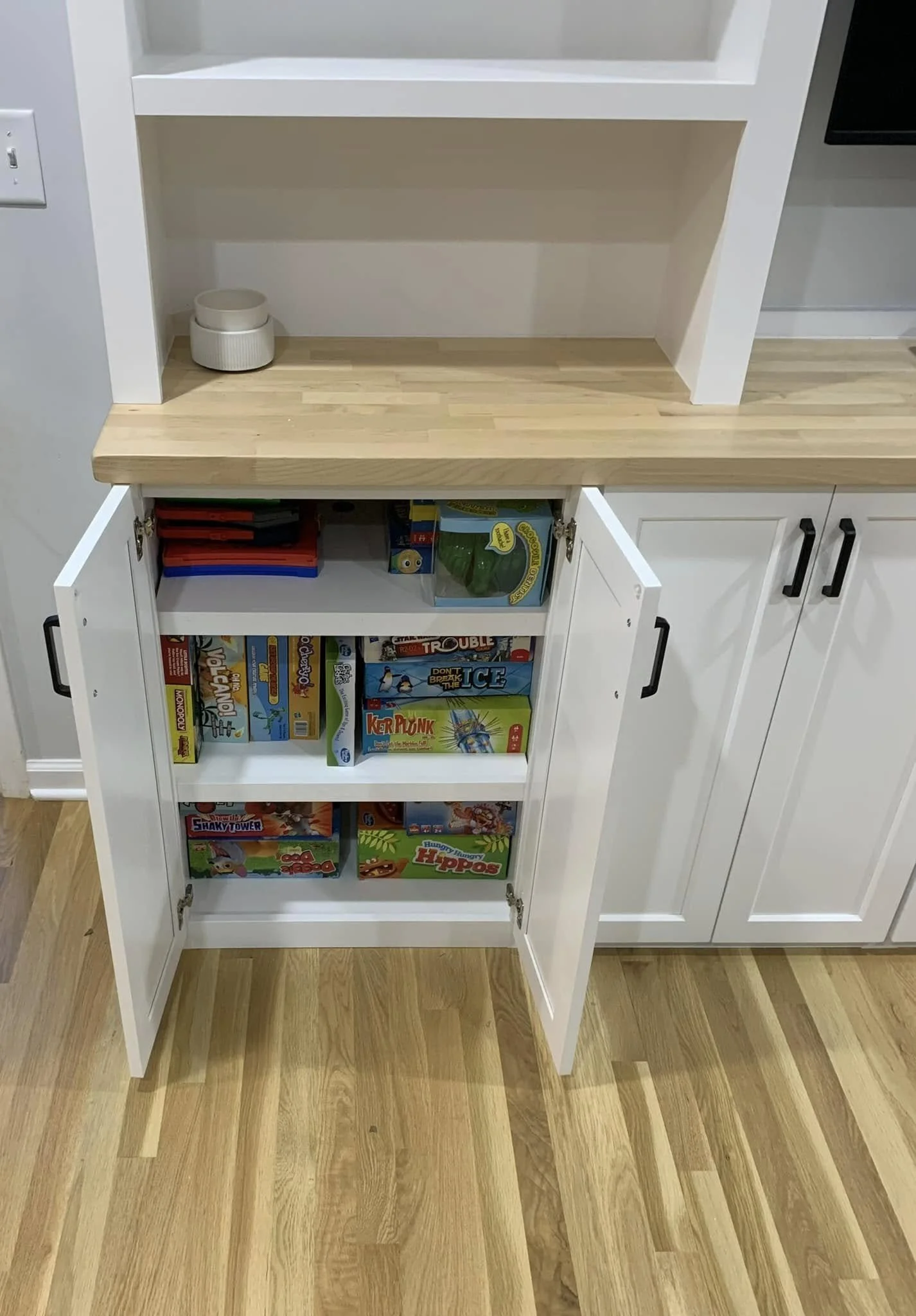 Open white kitchen cabinet with shelves containing board games and a wooden countertop above, and a small white container on the countertop, against a wall and wooden floor.