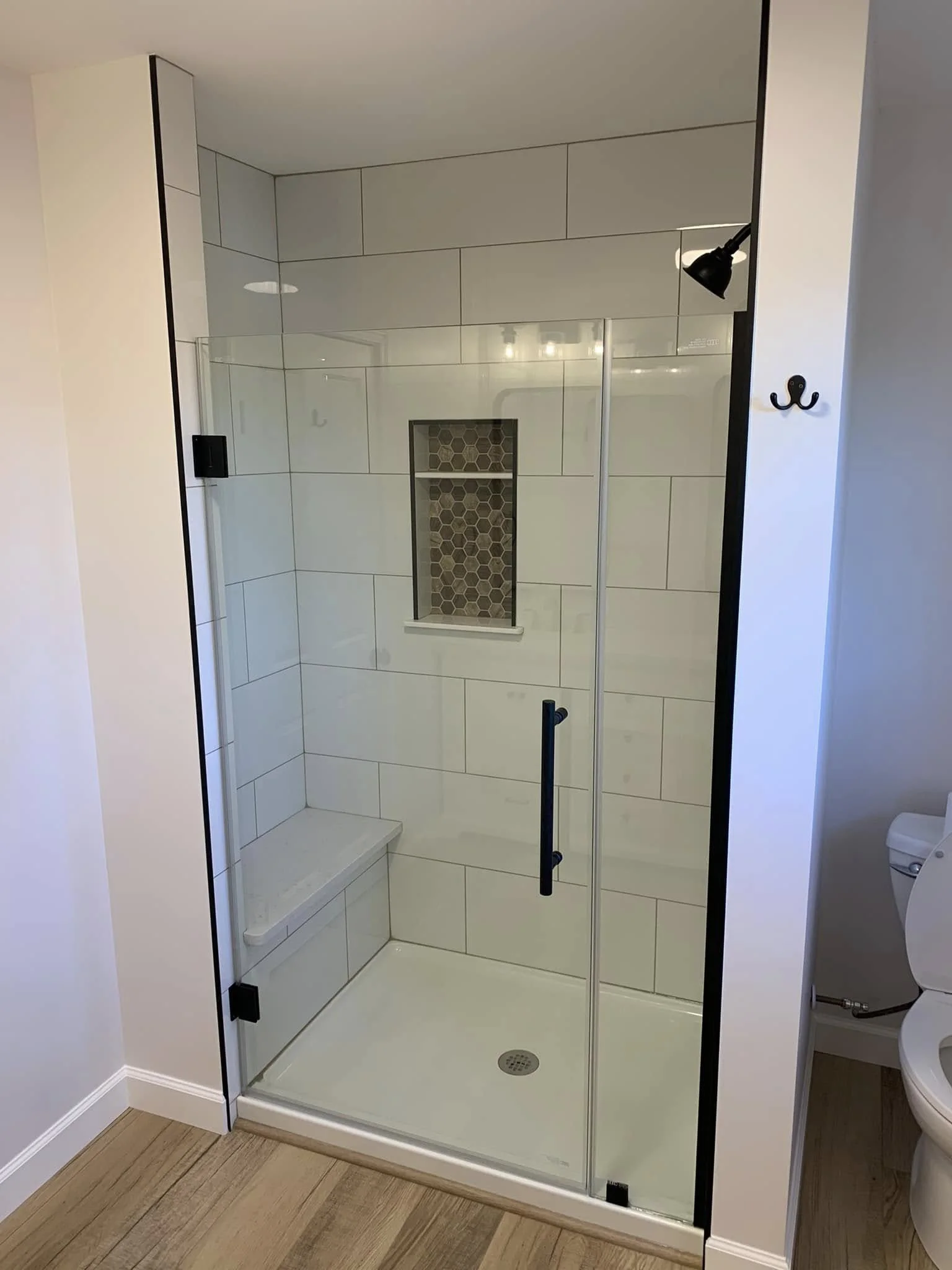 A modern walk-in shower with glass doors, white tiled walls, a small built-in shelf with hexagonal tiles, and a chrome showerhead against a white wall.