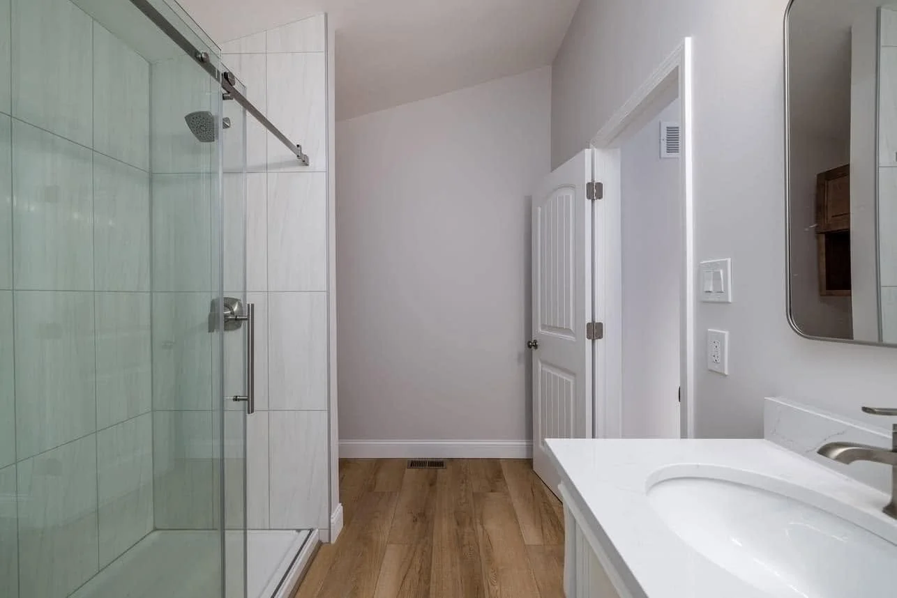 A bathroom with a glass-enclosed shower, a white vanity with a sink, a mirror, white walls, a wooden floor, and a partially open door.