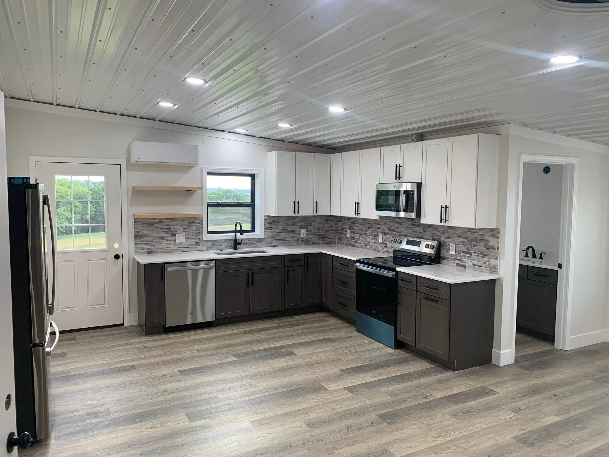 Modern kitchen with white upper cabinets, dark lower cabinets, a stainless steel oven, a microwave, and a tile backsplash. There are windows looking out to greenery, and a door leading outside. The ceiling has exposed metal panels and recessed lighti