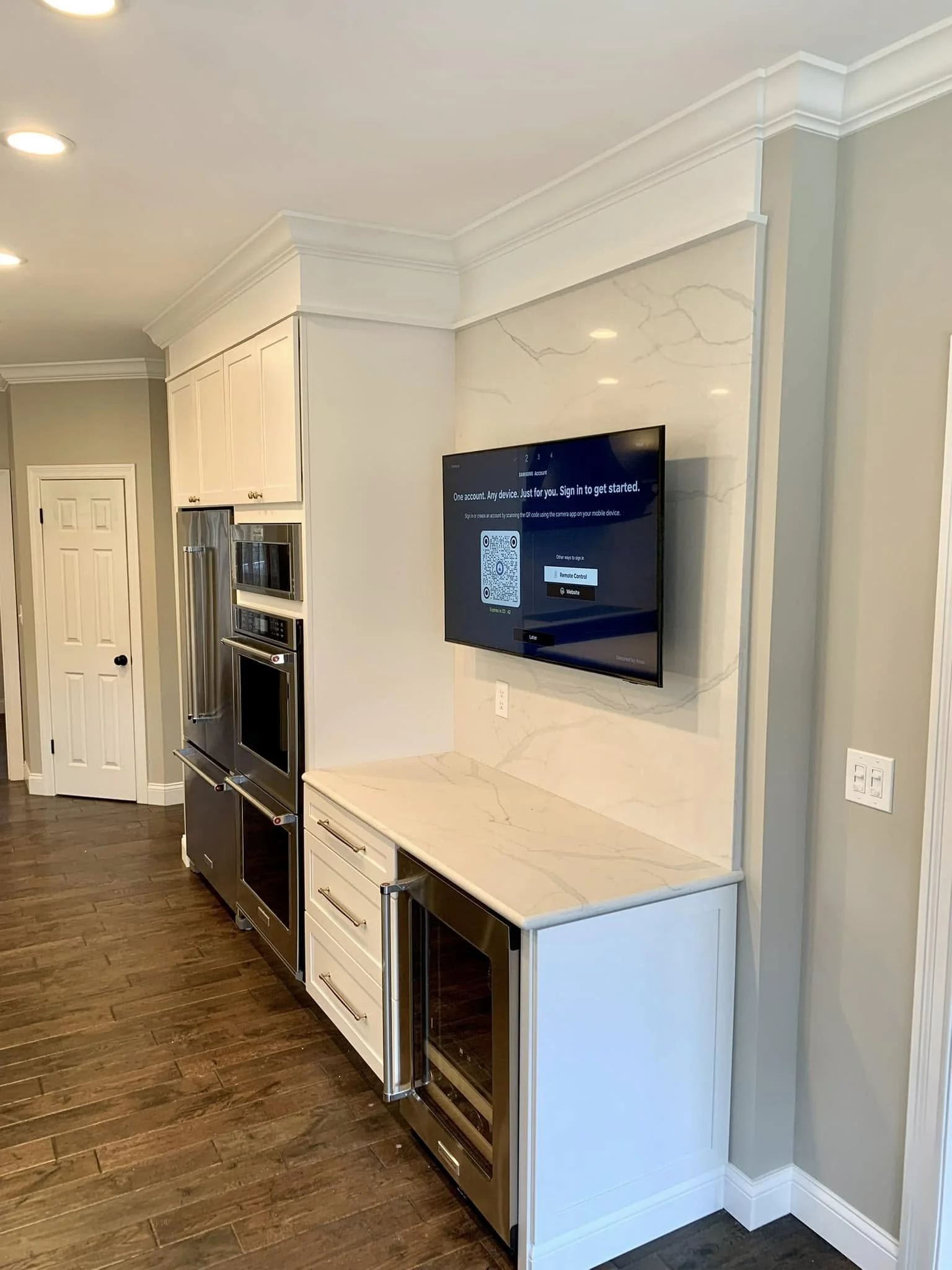 Kitchen area with built-in microwave, oven, and wine refrigerator, white cabinetry, marble countertop, and a mounted television on the wall.