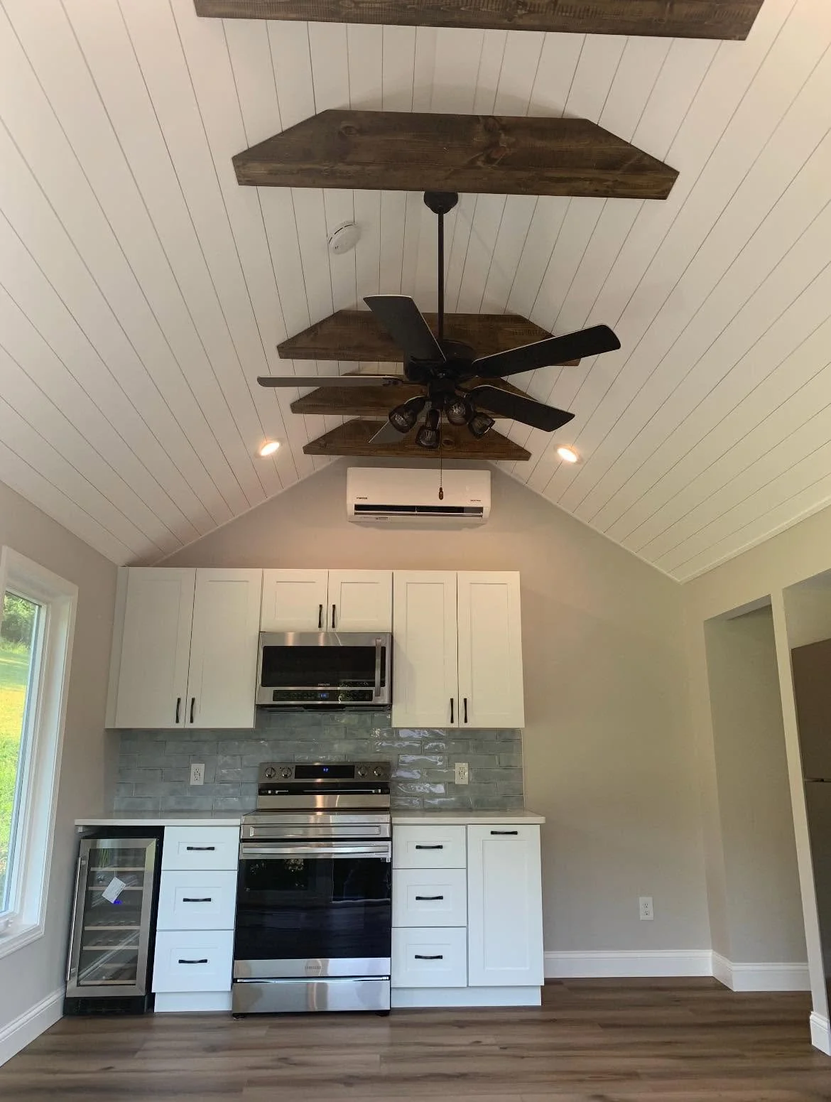 Kitchen with white cabinetry, stainless steel appliances, gray tiled backsplash, wood flooring, and a vaulted ceiling with three wooden shelves, a ceiling fan, and an air conditioning unit.