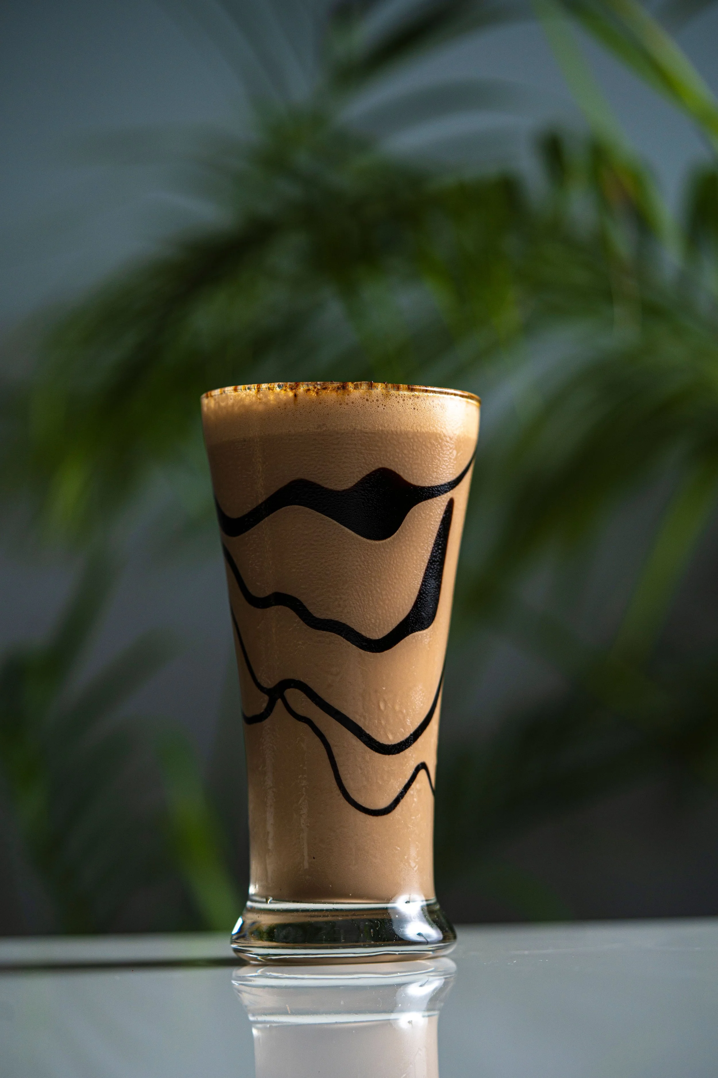 A glass of iced coffee with a latte art design on top, placed on a reflective surface with blurred green leaves in the background.