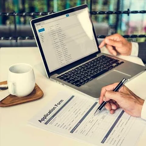 Person filling out an application form with a pen on a desk, with a laptop showing an email inbox open and a white mug nearby.