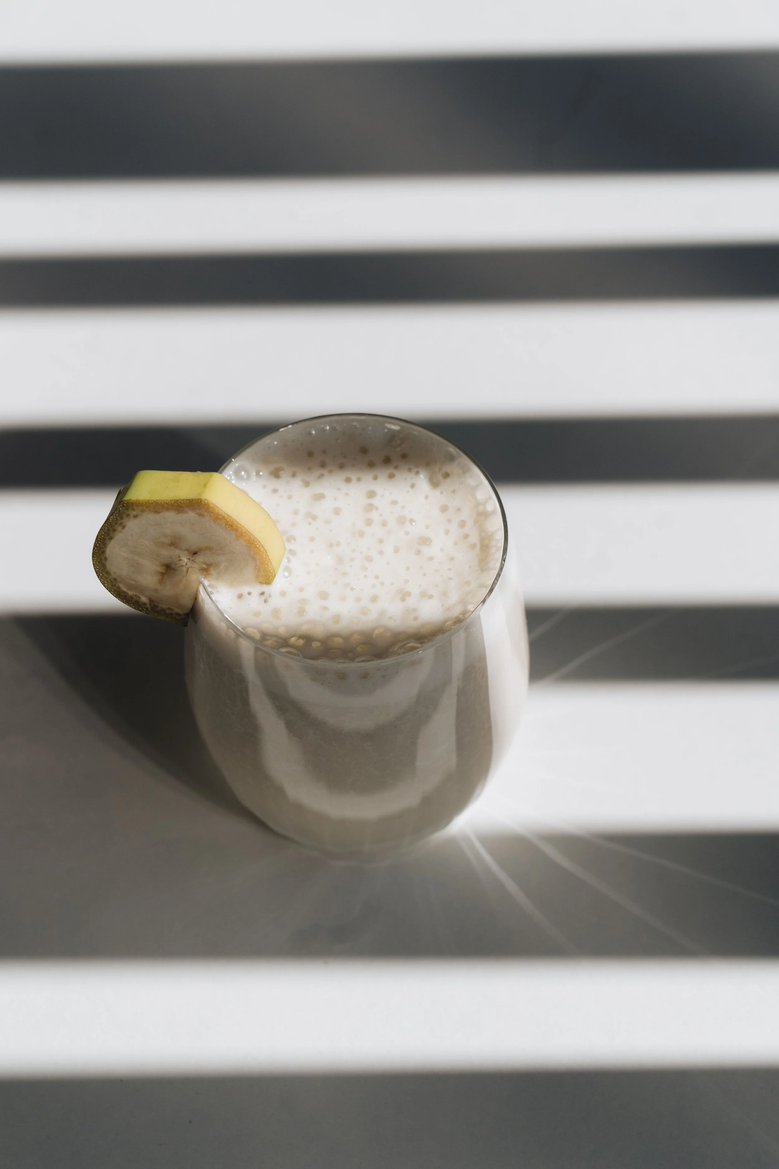 A glass of milk garnished with a slice of banana on the rim, placed on a white surface with shadow stripes in the background.
