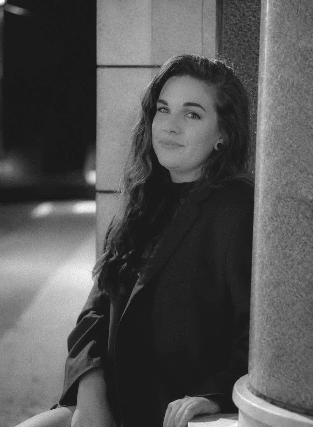 A woman with long wavy hair and dark clothing leaning against a wall in an urban setting, smiling at the camera.