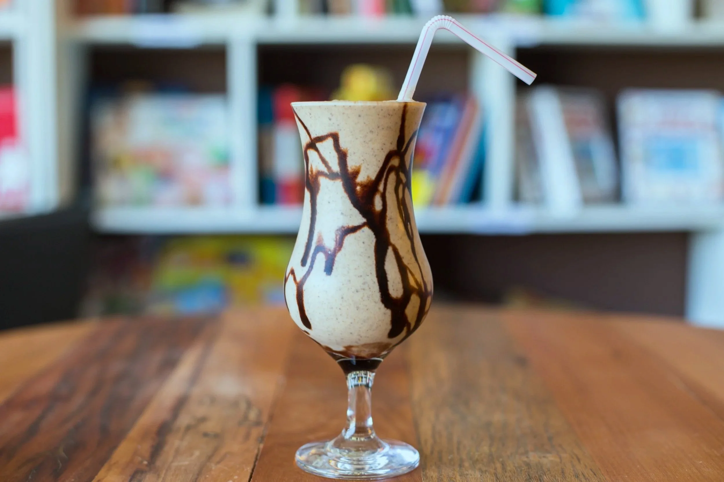 A tall glass filled with an ice cream milkshake, topped with chocolate syrup and a bent paper straw, on a wooden table with a blurred bookshelf in the background.