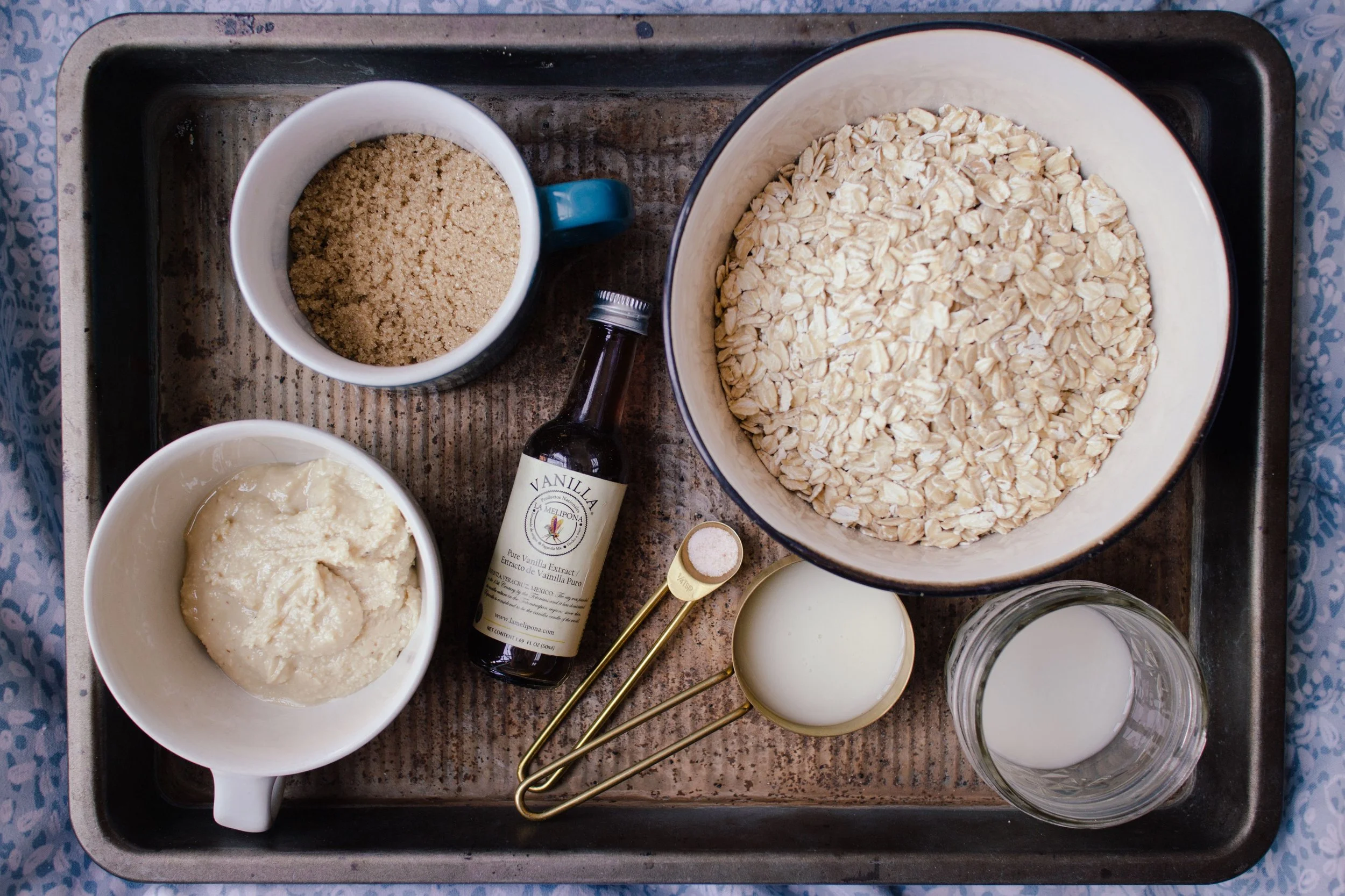 Overhead view of kitchen ingredients on baking tray including oats, brown sugar, vanilla extract, milk, a measuring spoon, and a bowl of cookie dough