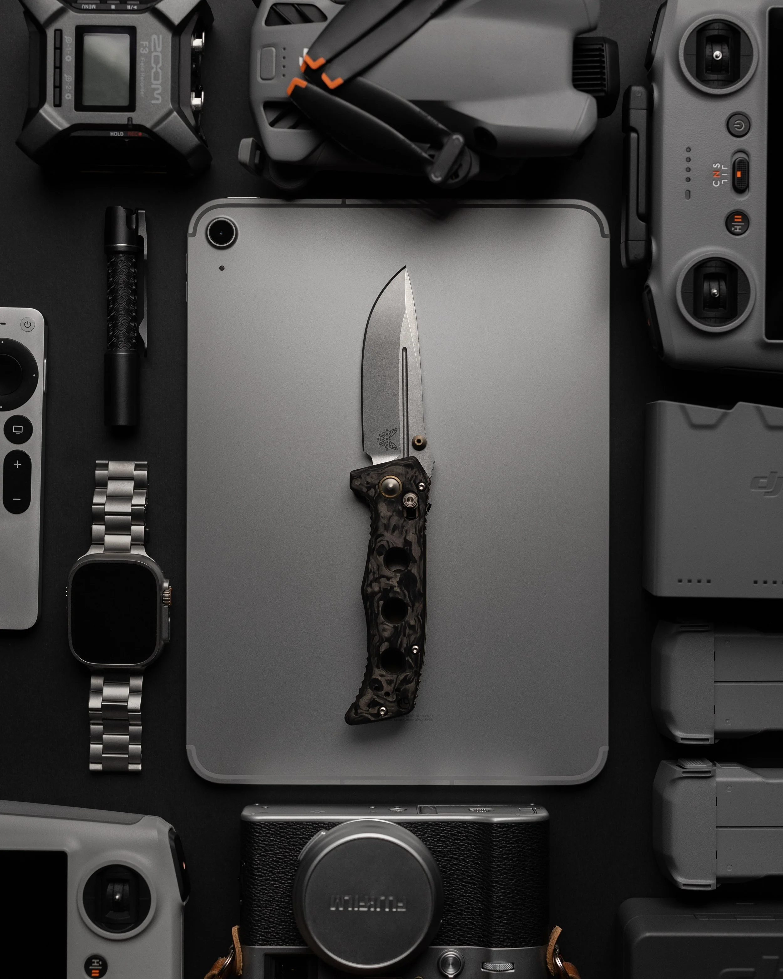 Top-down view of various photography equipment, including a camera, drone, smartphone, action camera, flash, wristwatch, and pocket knife, arranged on a dark surface.