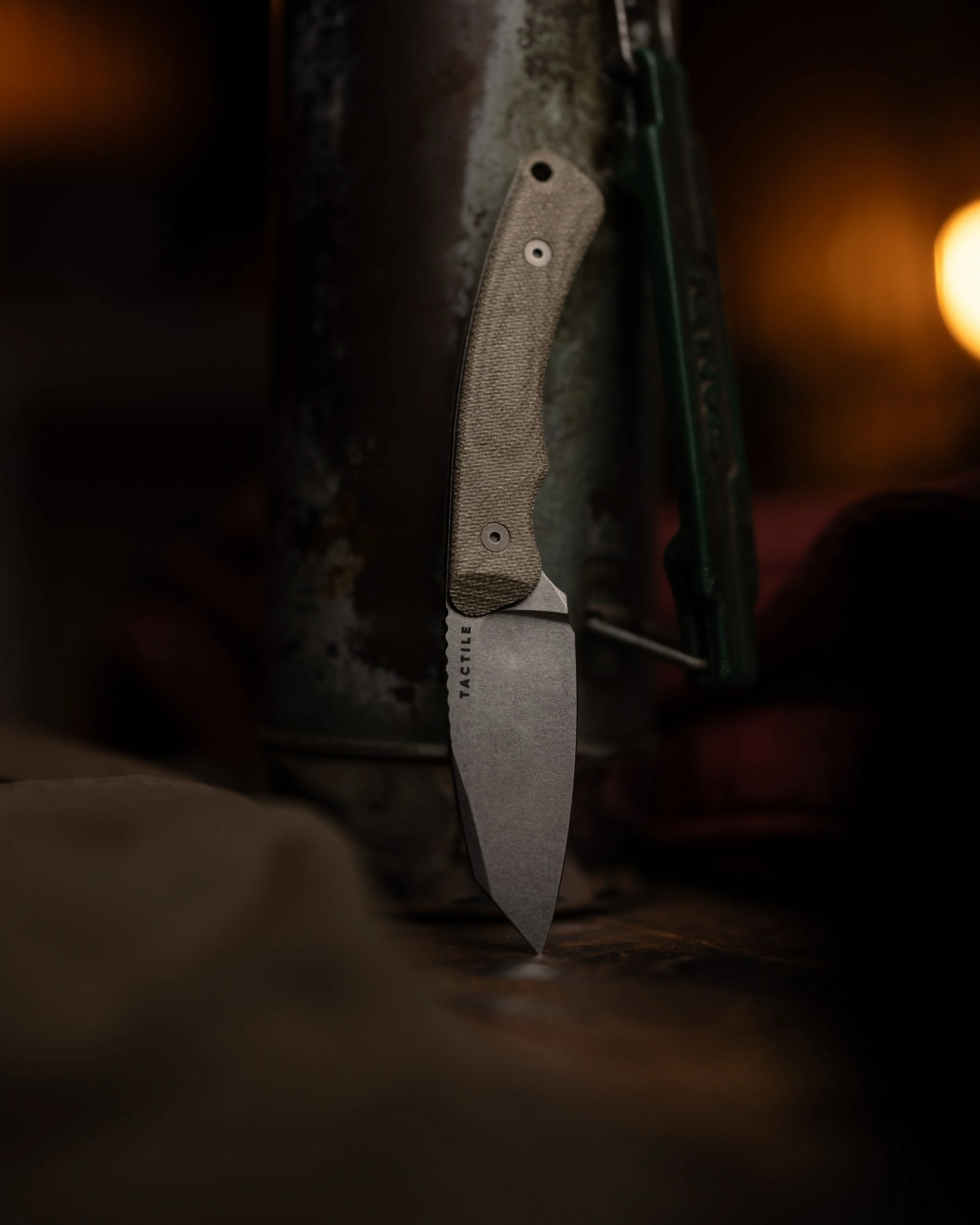 A close-up view of a Folding Tactile knife with a textured gray handle, partially hidden behind a dark surface, with warm orange lighting creating a blurred background.