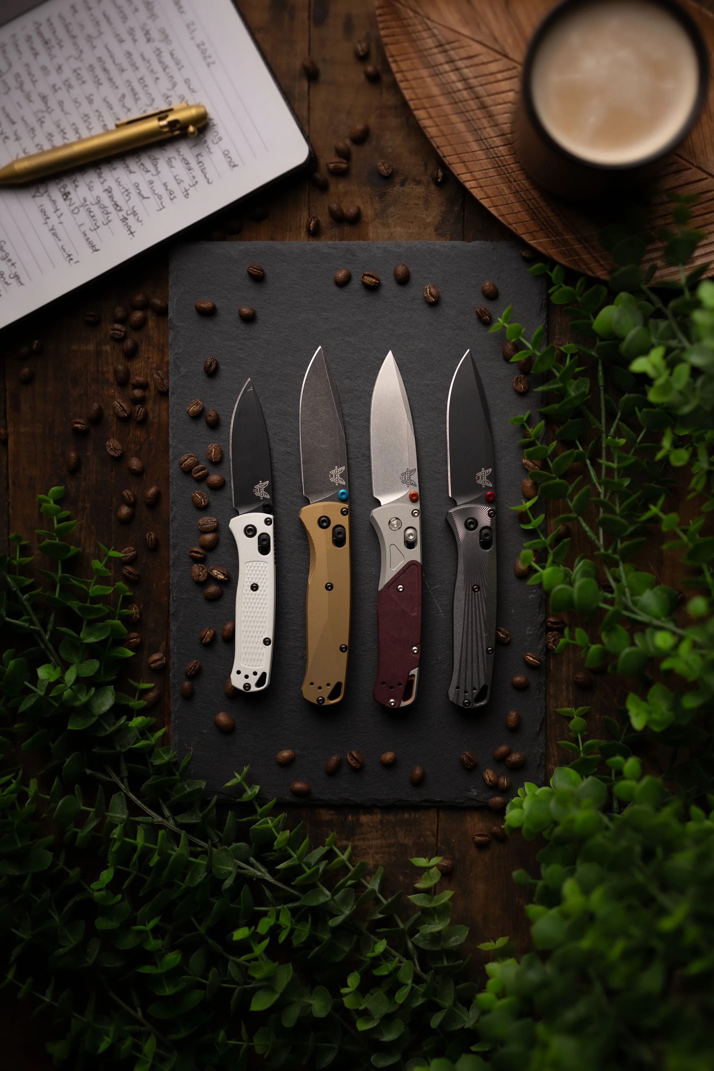 Four folding knives with different colored handles lie on a black slate board surrounded by scattered coffee beans and green foliage. A wooden tray with a beverage in a dark mug and a notebook with a gold pen are also visible.