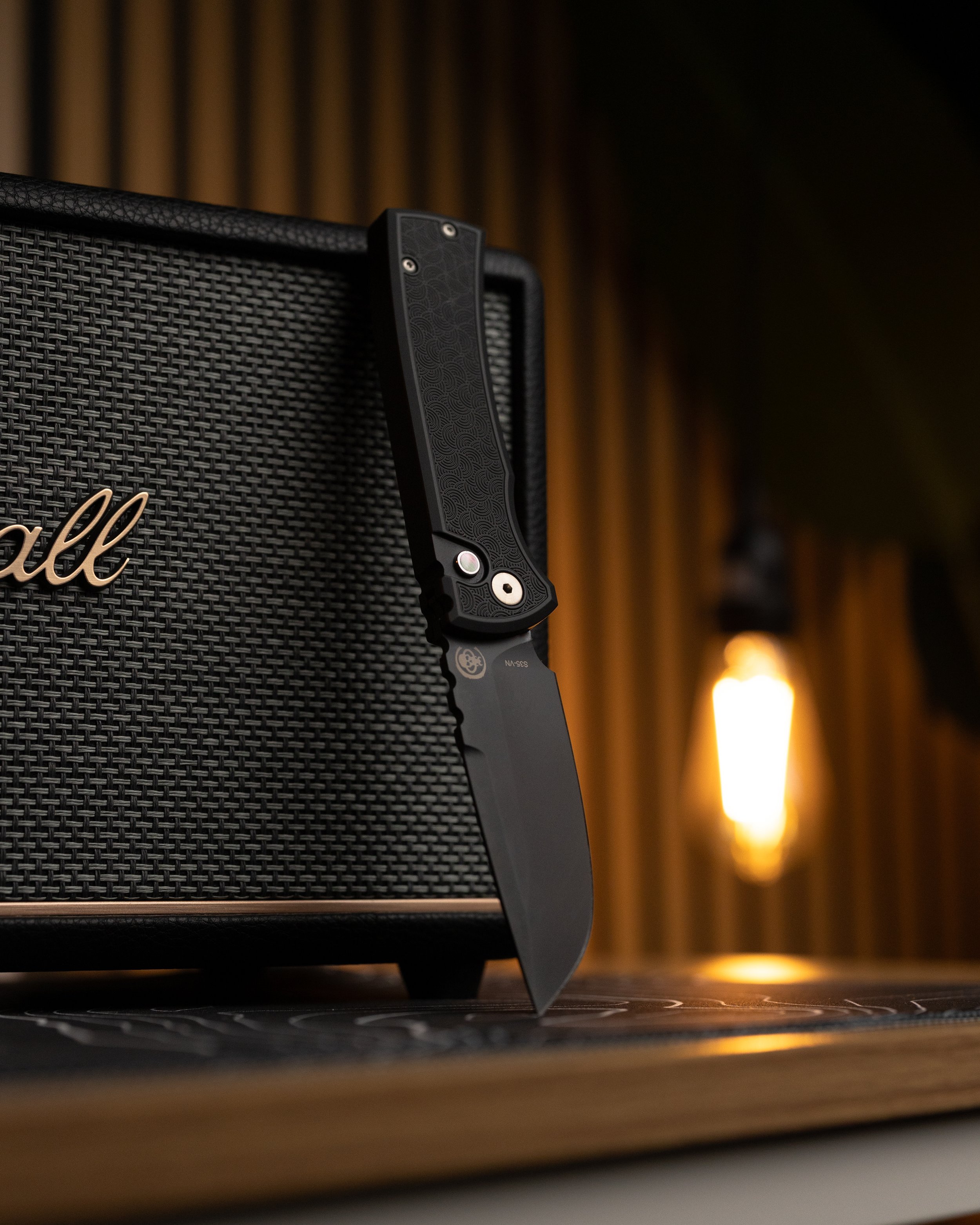 Close-up of a black folding pocket knife with a textured handle, positioned upright against a black textured speaker or amplifier with the brand name partially visible. The background includes warm yellow lights and a blurred wooden wall.