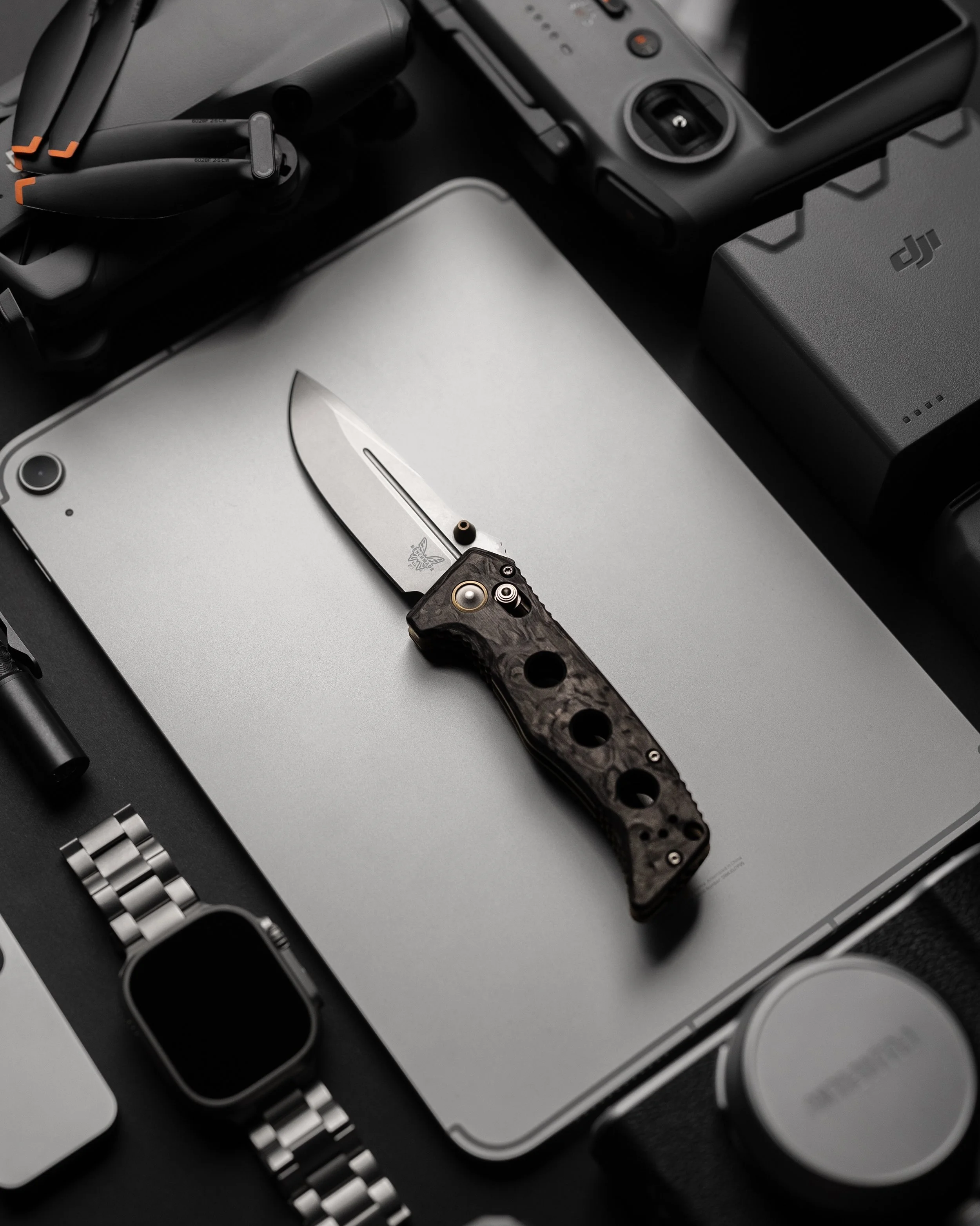 A collection of electronic gadgets and tools including a DJI drone controller, a folding knife, a camera, a smartwatch, and a drone battery, arranged on a metallic surface.