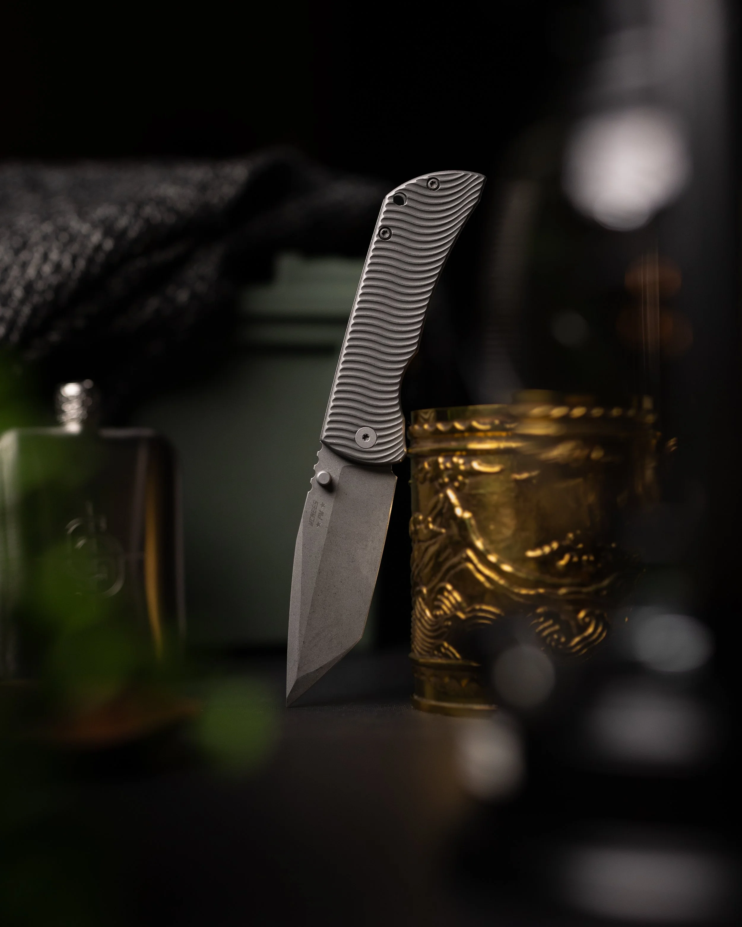 A partially opened folding knife with a textured handle beside a gold-colored decorative container, with blurred objects in the background.