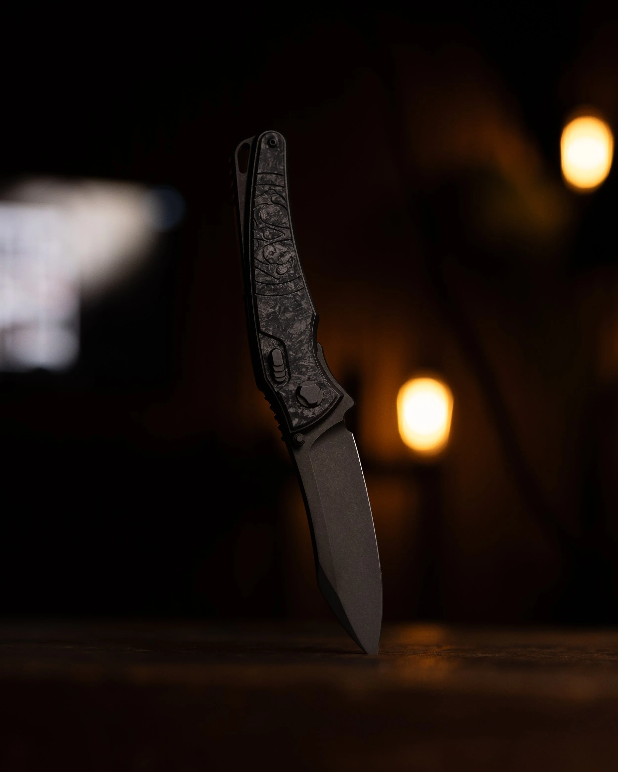 Close-up of a folding knife with a patterned handle, displayed against a dark background with warm bokeh lights.