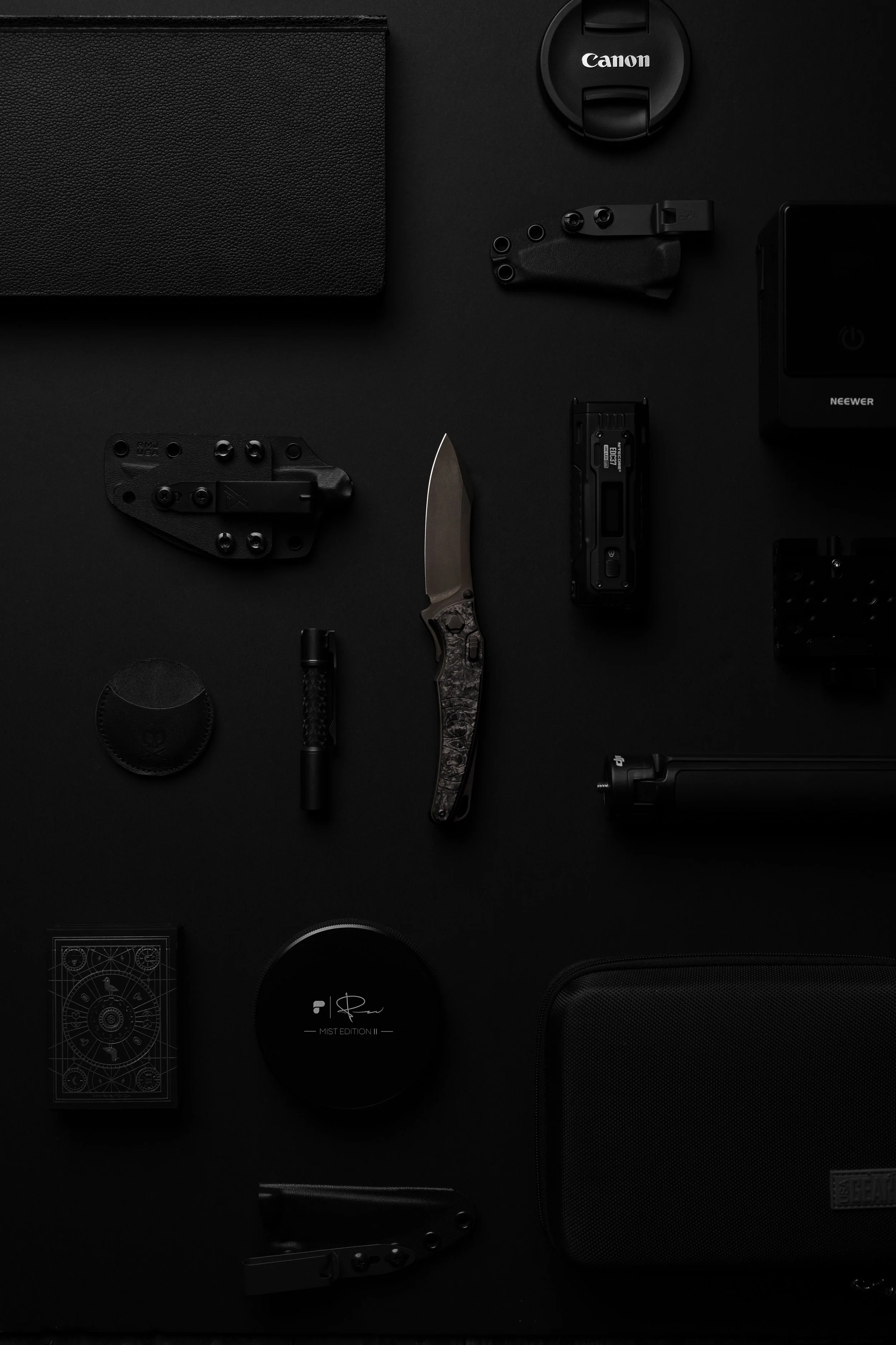 Collection of black tech gadgets and accessories arranged on a dark surface, including a camera lens cap, a small knife, a power bank, a portable speaker, and various electronic devices and cases.