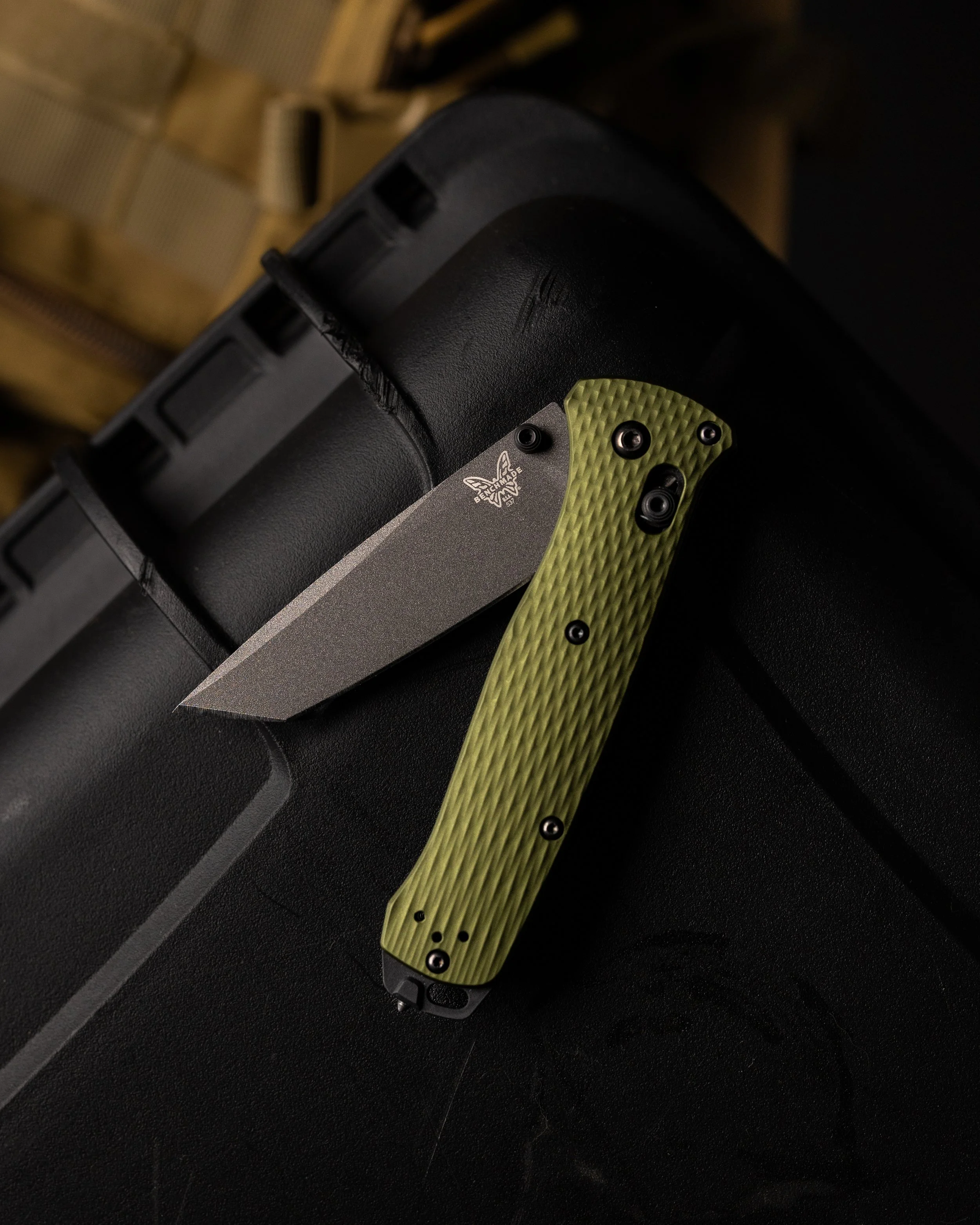Close-up of a folding knife with a black handle and a metallic blade, resting on a black surface. The handle has a green textured grip and black screws. The blade has a logo and text reading a butterfly logo and 'BENCHMADE'.