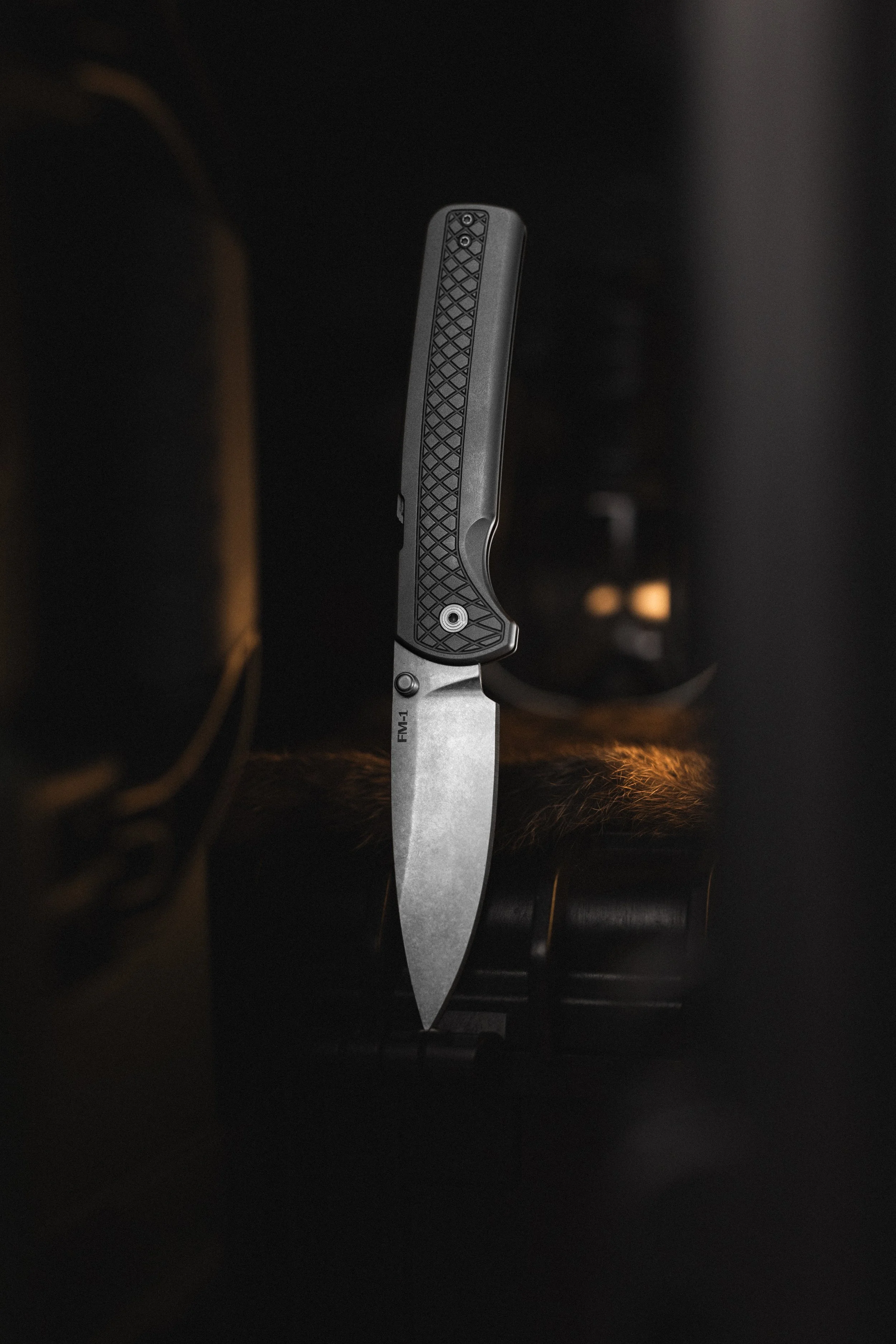 A close-up photograph of a knife with a textured grip, standing upright on a dark surface with a black background.