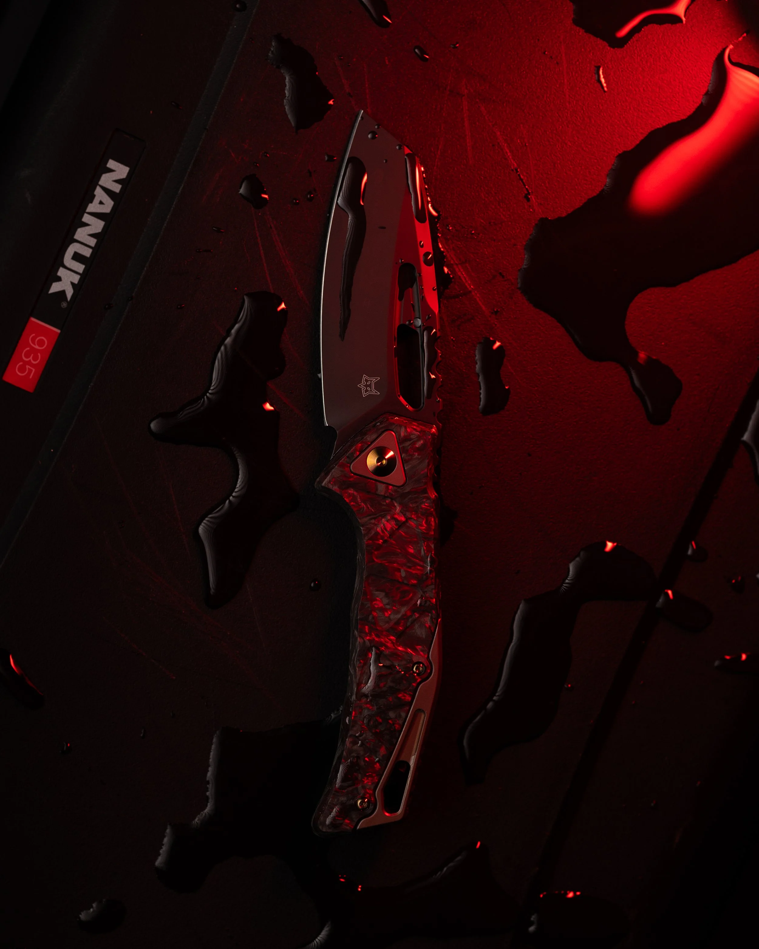 A knife with a black and red handle and a gray blade with logo, resting on a dark surface with red lighting, surrounded by splashes of liquid.