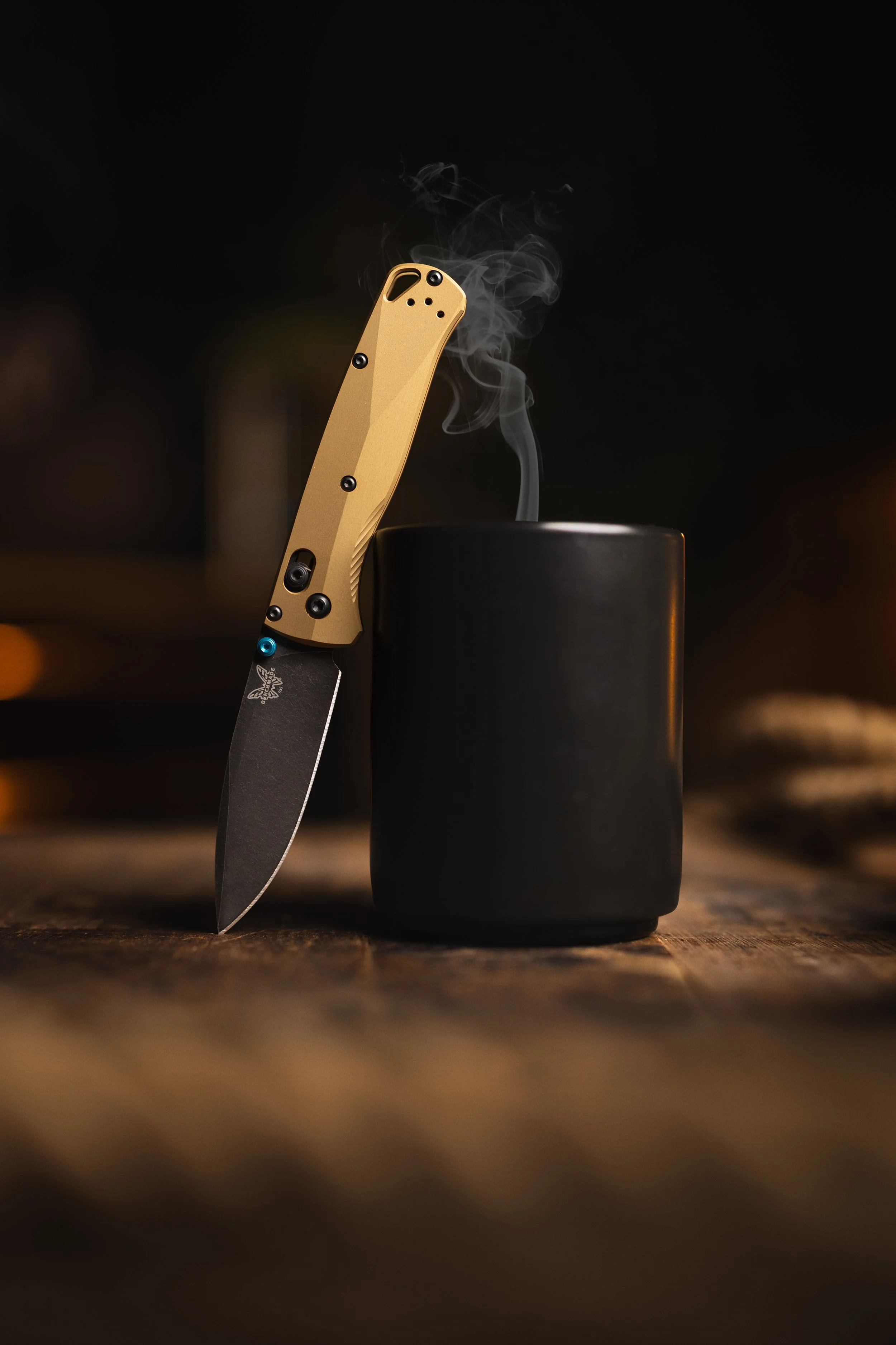 A folding knife with a tan handle and black blade, placed upright on a wooden surface, next to a black container with vapor rising from it, against a dark background.