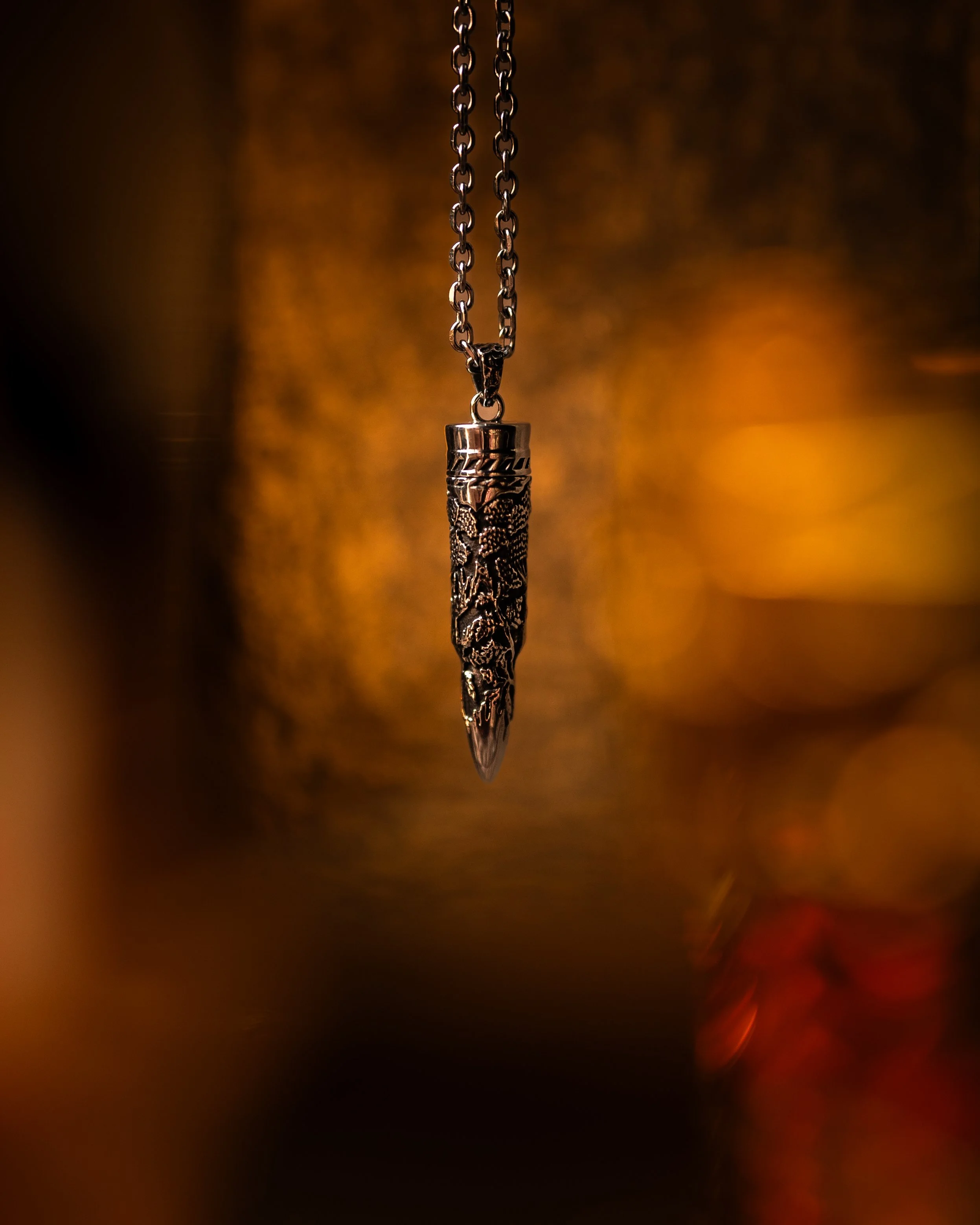 Close-up of a silver pendant necklace with a detailed, textured design hanging from a chain against a blurred, warm background.