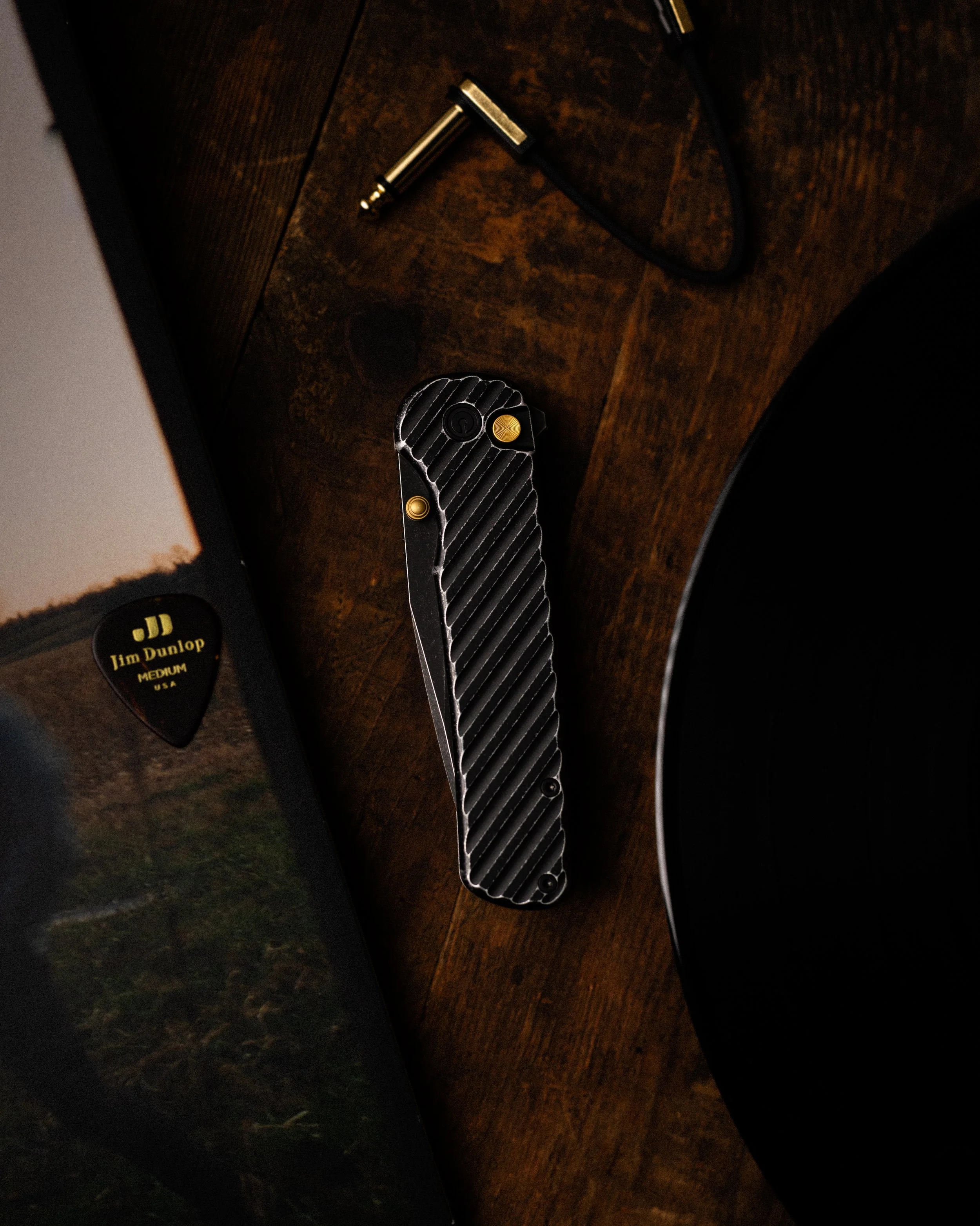A flat metal tool with a textured grip, a black and yellow cable, a partially visible Jim Dunlop guitar pick, and a small black and yellow box on a dark wooden surface.