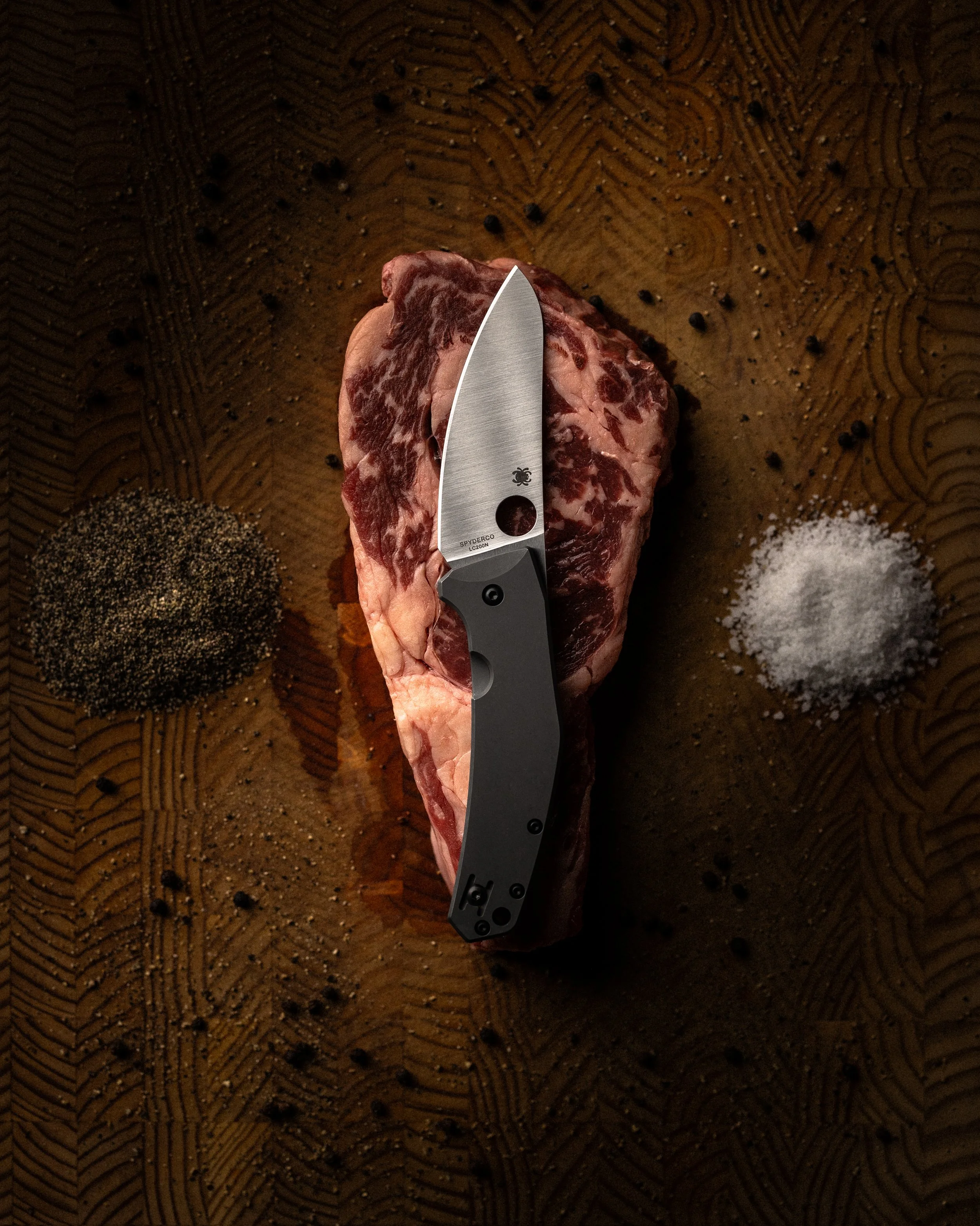 A piece of marbled raw steak on a wooden cutting board with black pepper, salt, and ground spices around it. A gray pocket knife rests on the steak.
