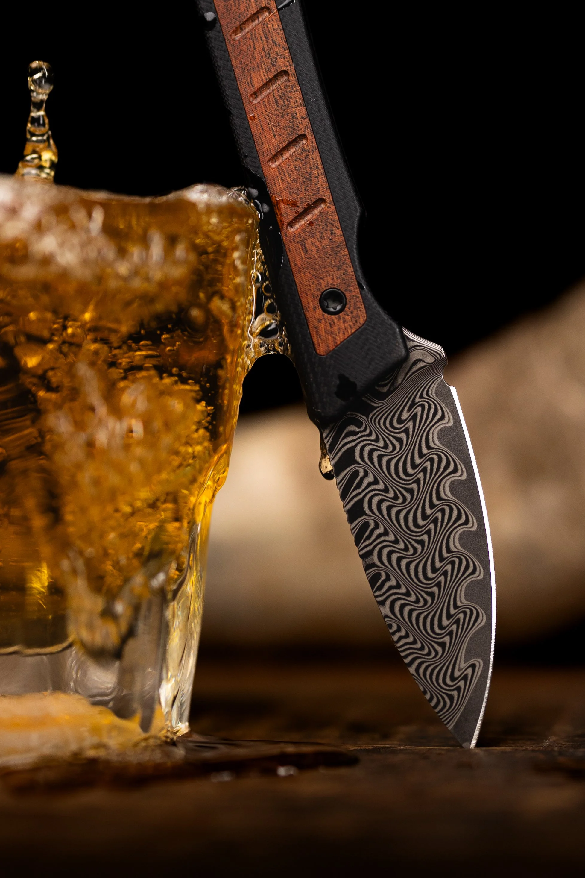 Close-up of a kitchen or combat knife with a patterned blade and a textured handle, inserted into a glass of beer with bubbles and foam near the surface, on a wooden surface against a dark background.
