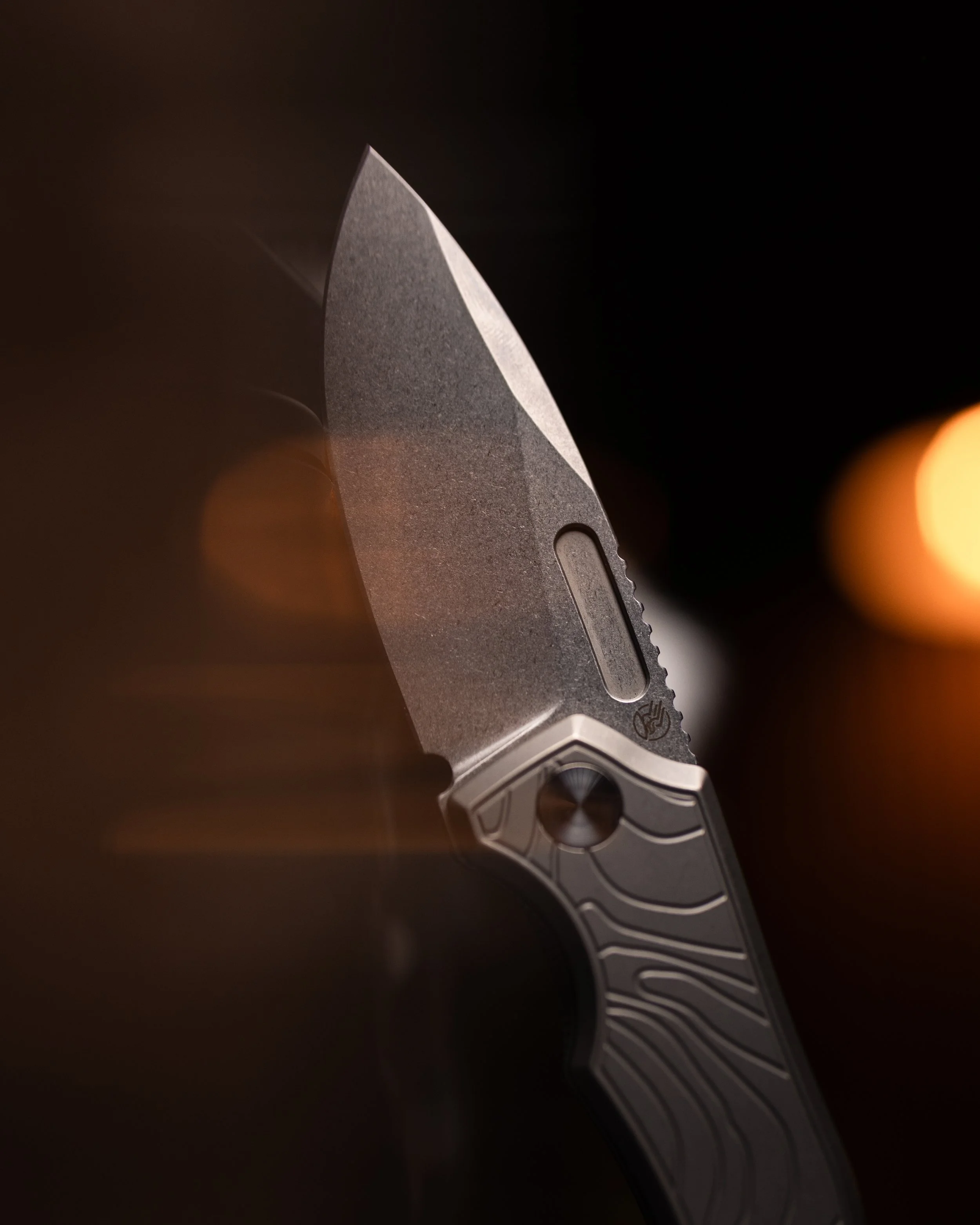 Close-up of a metallic folding knife blade with a textured surface, partially open, with a pattern engraved on the handle, set against a dark background with warm light flares.